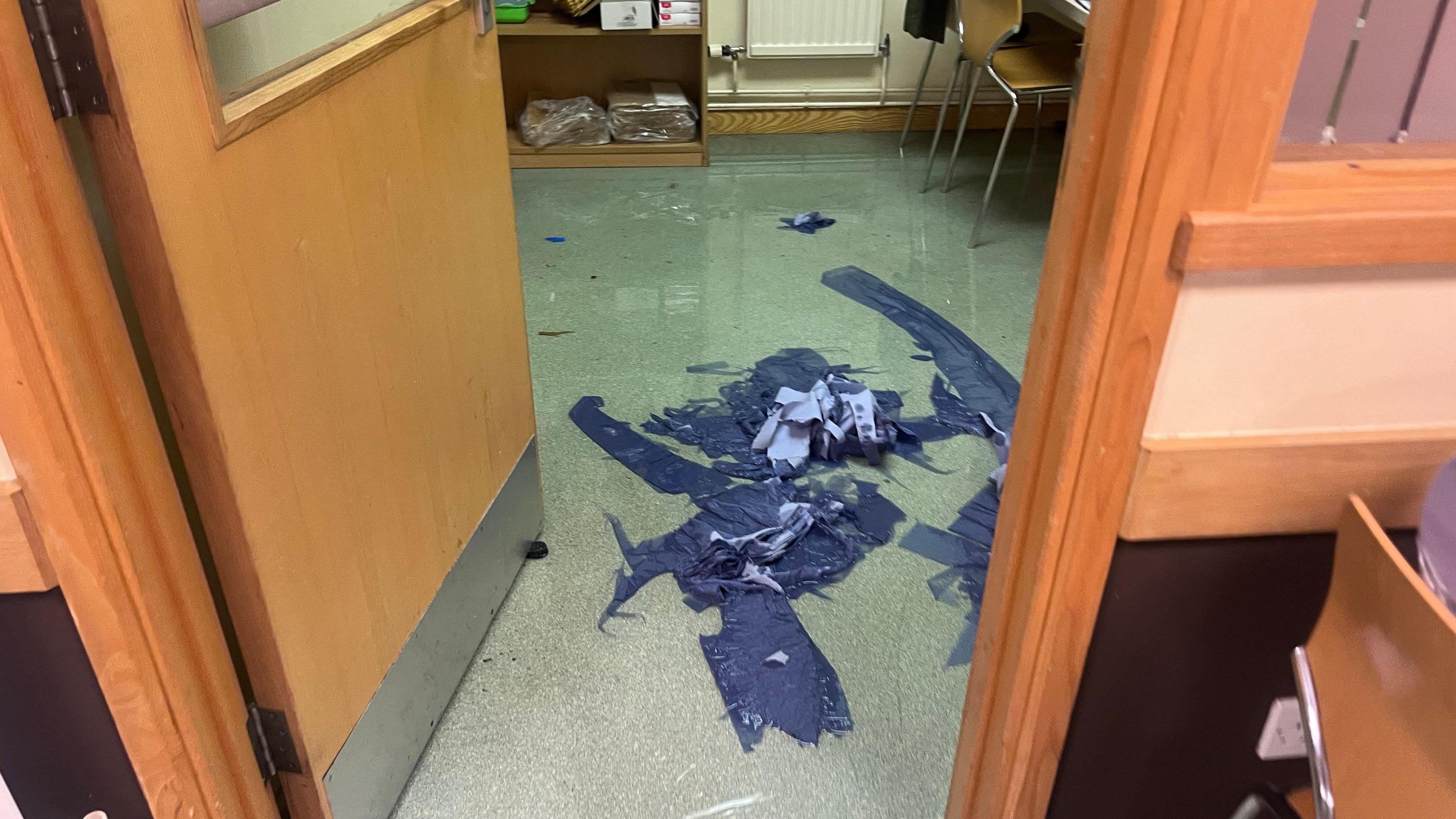 Floodwater can be seen covering the floor, with a heap of sodden blue paper visible in the centre of the room where people have tried to clean the flood water. 