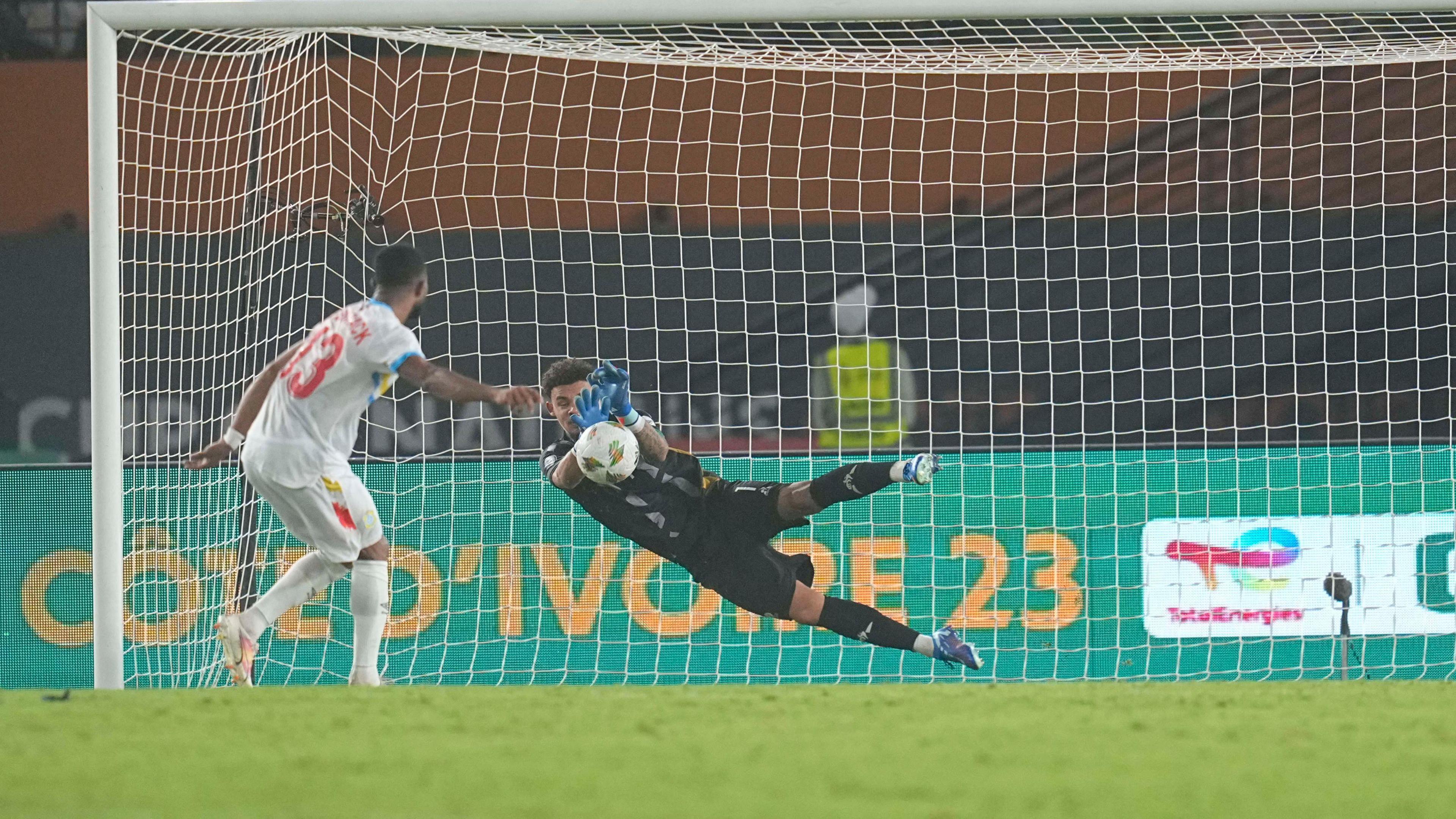 Ronwen Williams in black goalkeeping kit dives to his right and raises his blue gloves with palms facing forwards to save a ball that has been sent goalwards from the penalty spot by a DR Congo player during the third-place play-off at the 2023 Africa Cup of Nations. The picture is taken from around the halfway line, looking back towards the goal, meaning the Congolese player, in white kit, is seen from behind having just taken his spot-kick