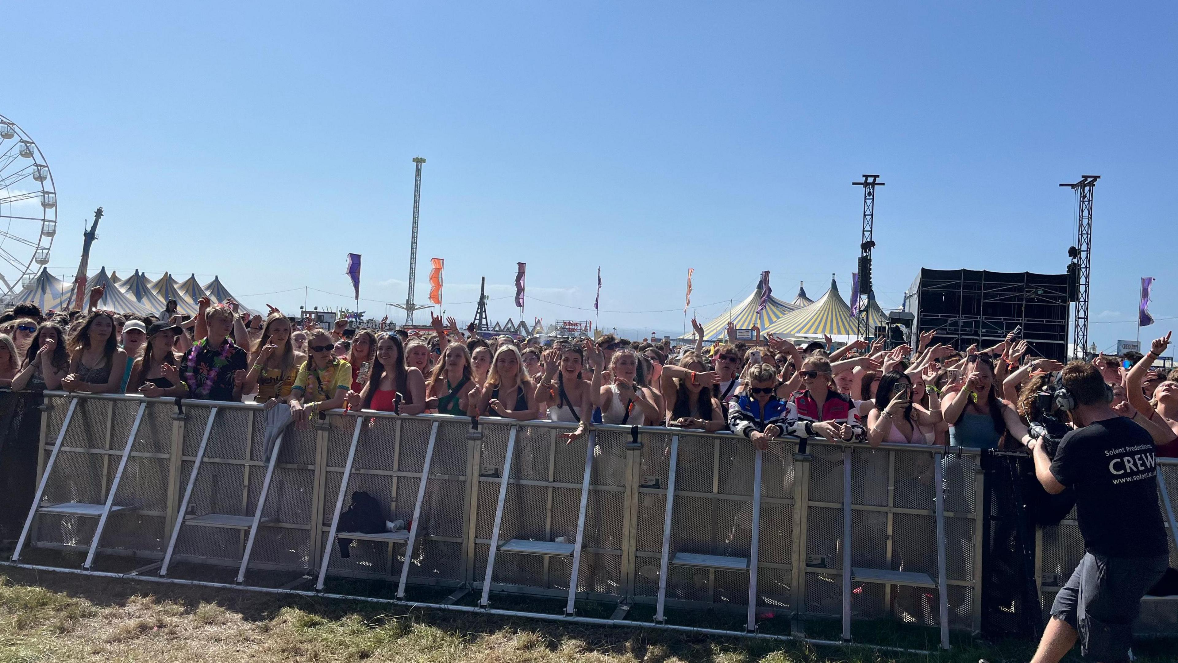 Thousands of people stand behind a barrier at Boardmasters in Cornwall. A camera man is on the right of the image. A big top and ferris wheel are in the background.