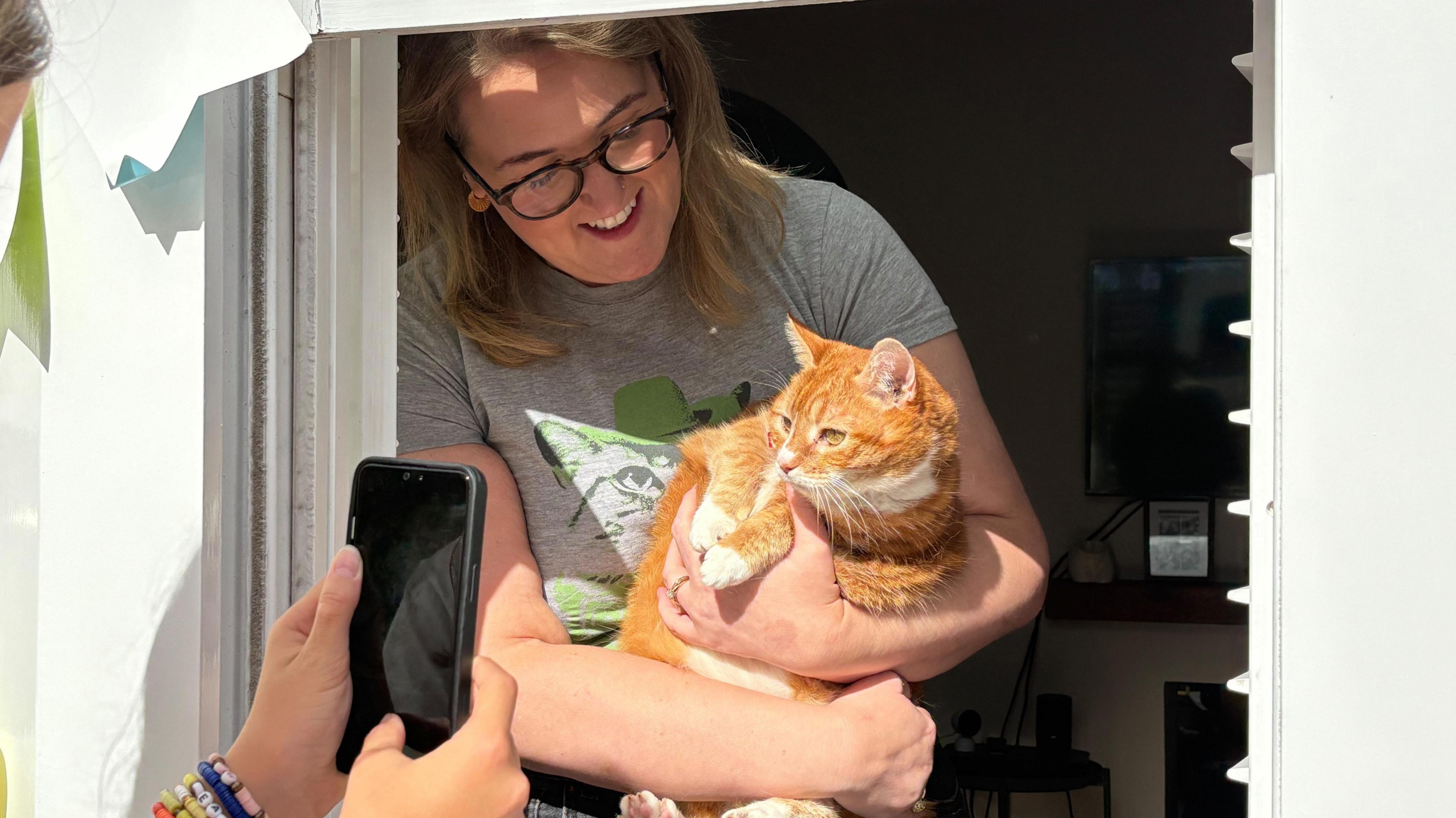 A woman behind an open window holds her ginger cat, Molly, while a passing visitor takes a close-up photo using her iPhone. The cat appears to be looking directly into the lens of the phone camera, with the owner smiling while looking down at her.