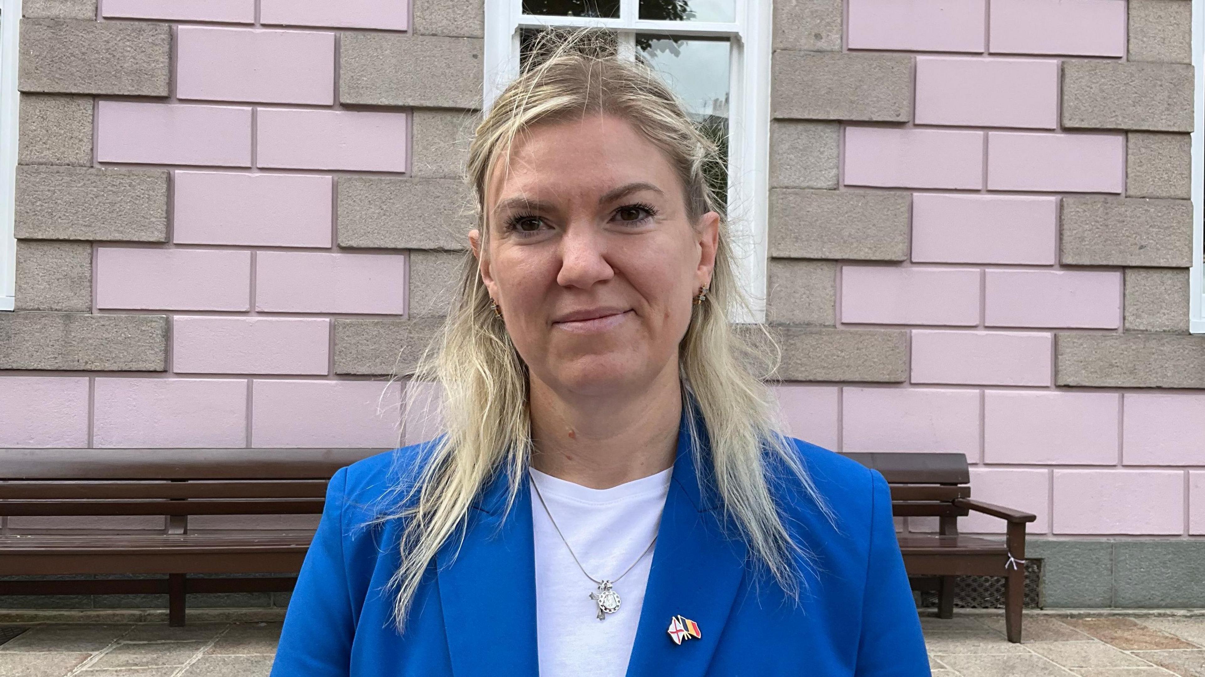 Deputy Raluca Kovacs stands outside a pink building. She has a blue blazer on and a white t-shirt underneath. She has a badge on her left lapel with the Jersey and Belgium flags on it. She has shoulder-length blonde hair.