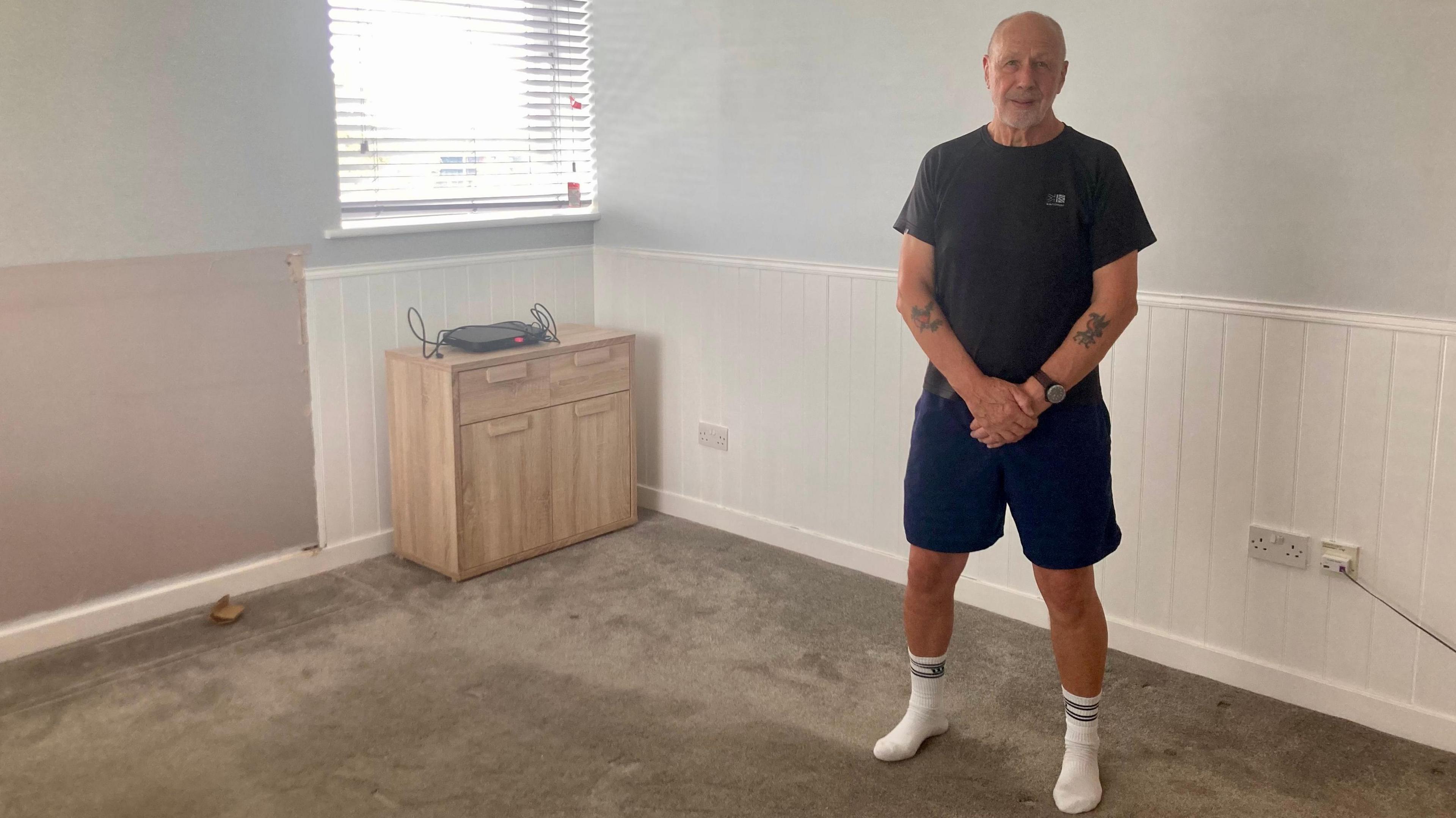 Dave Hemmings, who is bald and has a short beard, stands in his empty flat. There is only a carpet and a small cupboard left in the bare space.