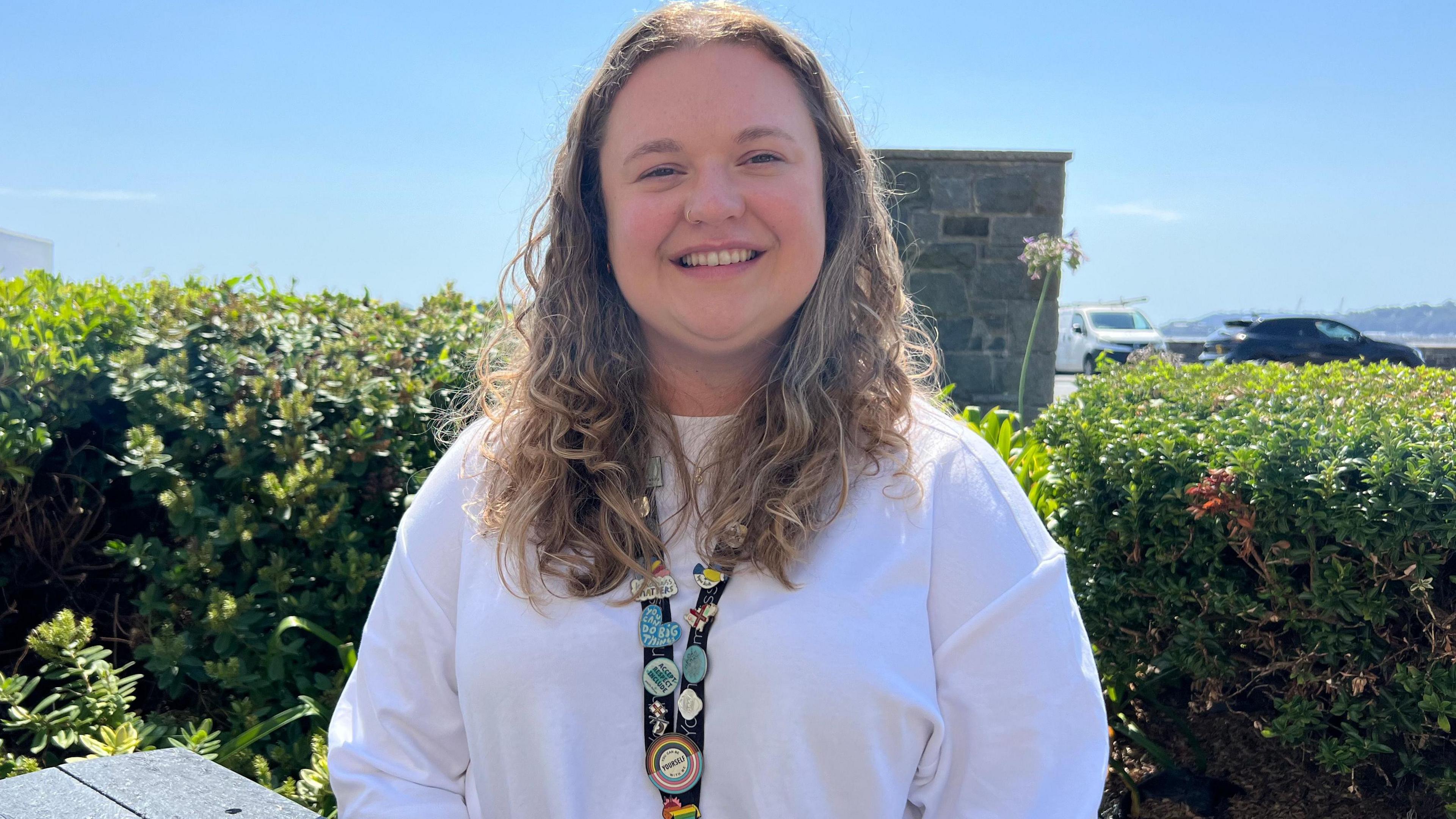 Maisie Foote has shoulder length hair and is wearing a white T-shirt and a lanyard with a lot of differently shaped and coloured badges on it. She is smiling and standing in front of a green hedge on a sunny day.