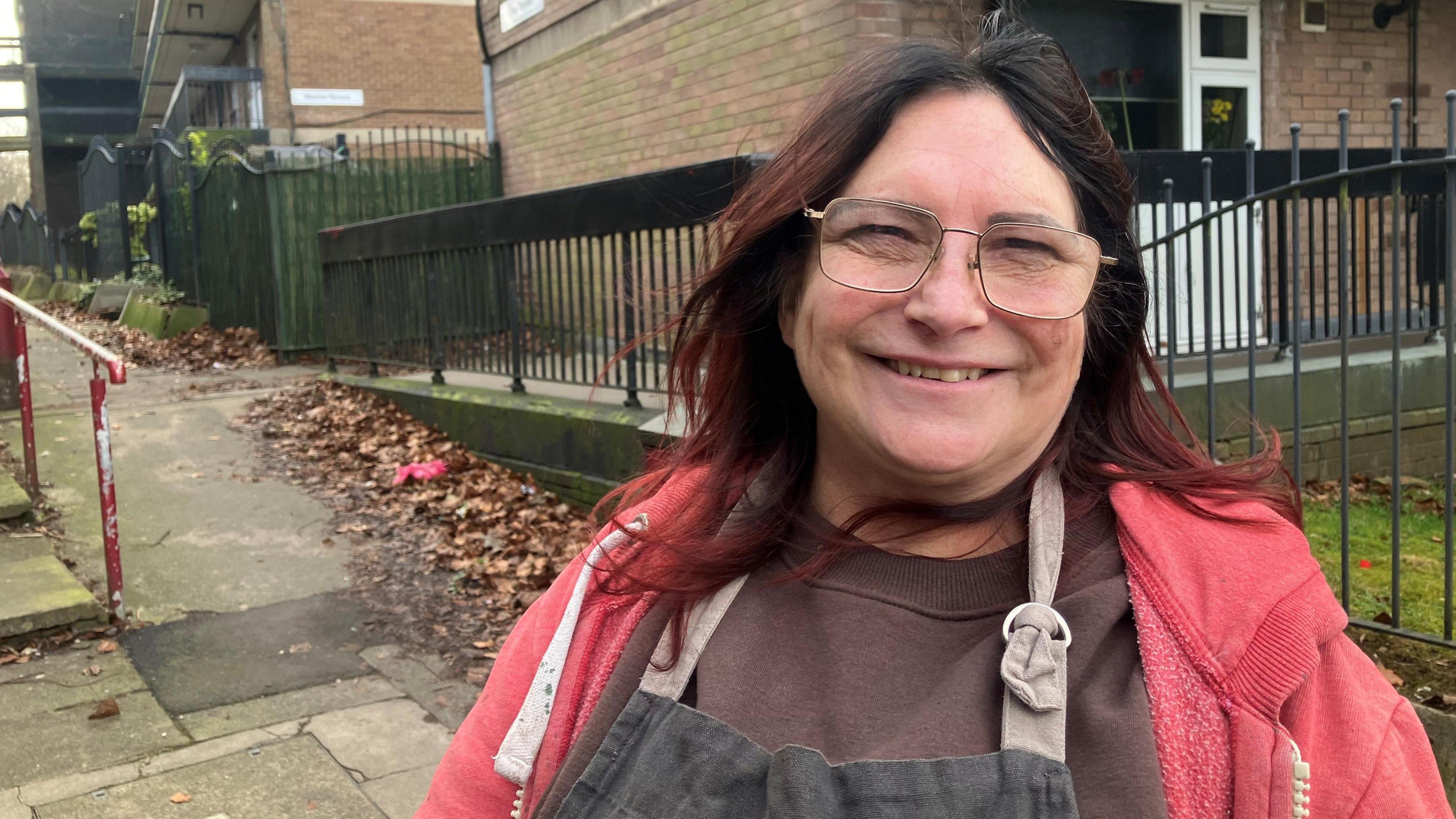 Lorraine Simpson, 61, who has shoulder length dark red hair and wears glasses, smiles at the camera against the backdrop of a three-storey maisonette 