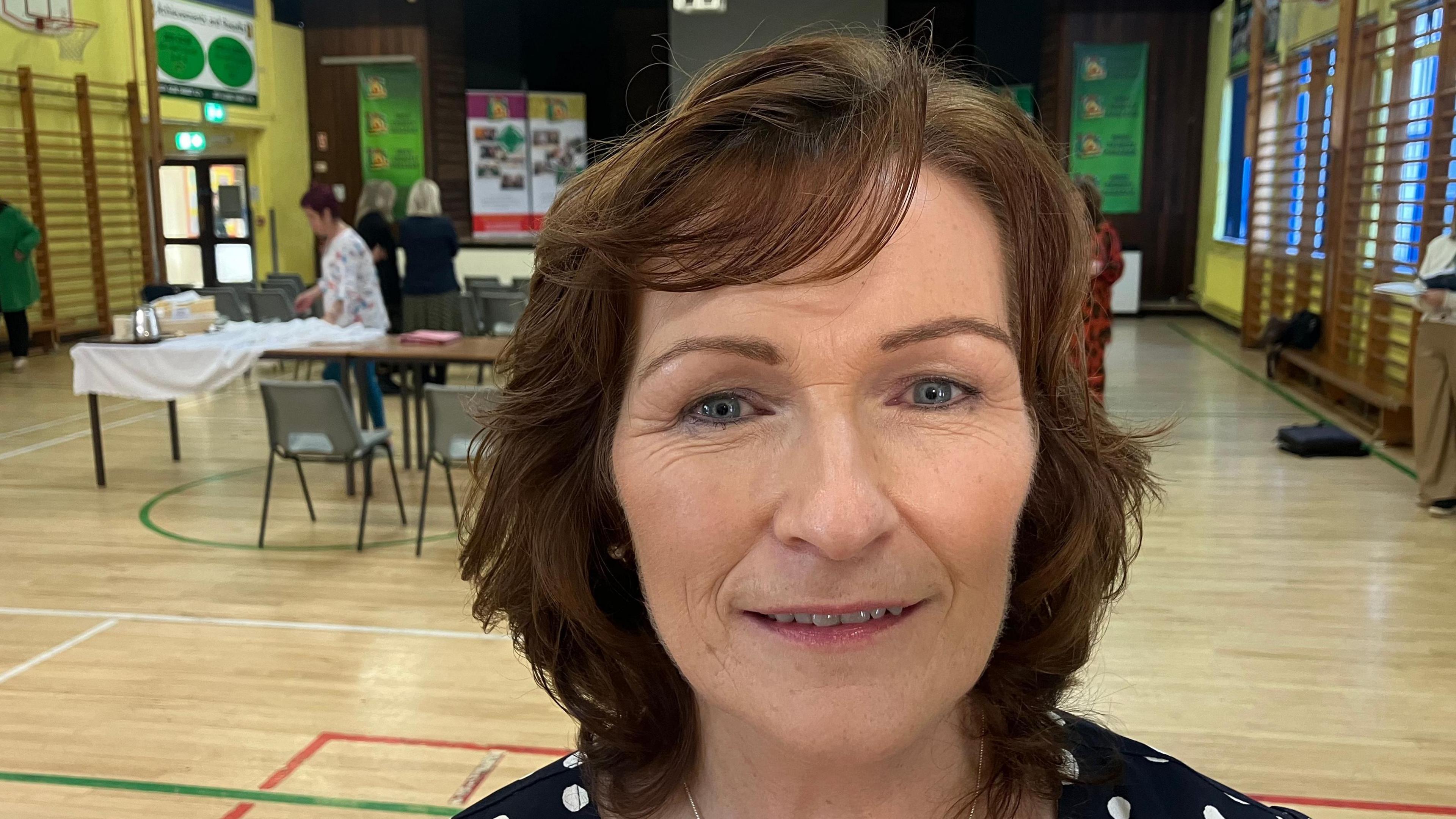 Photo of Isabel Russell who has brown short hair and is wearing a black and white polka dot top standing in a school sports hall