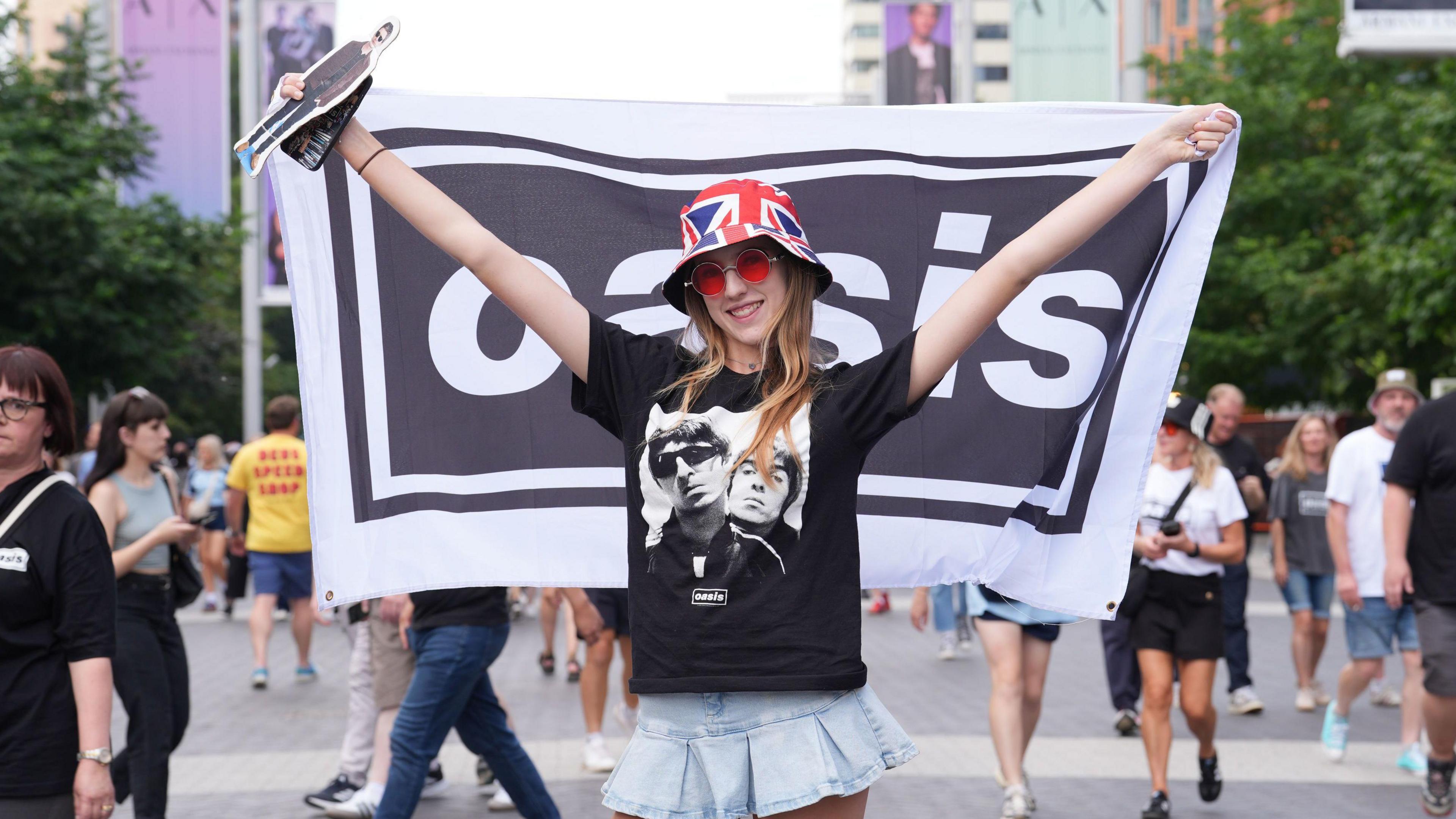 A woman wearing a black t-shirt featuring Noel and Liam Gallagher holds a flag with the Oasis band logo. 