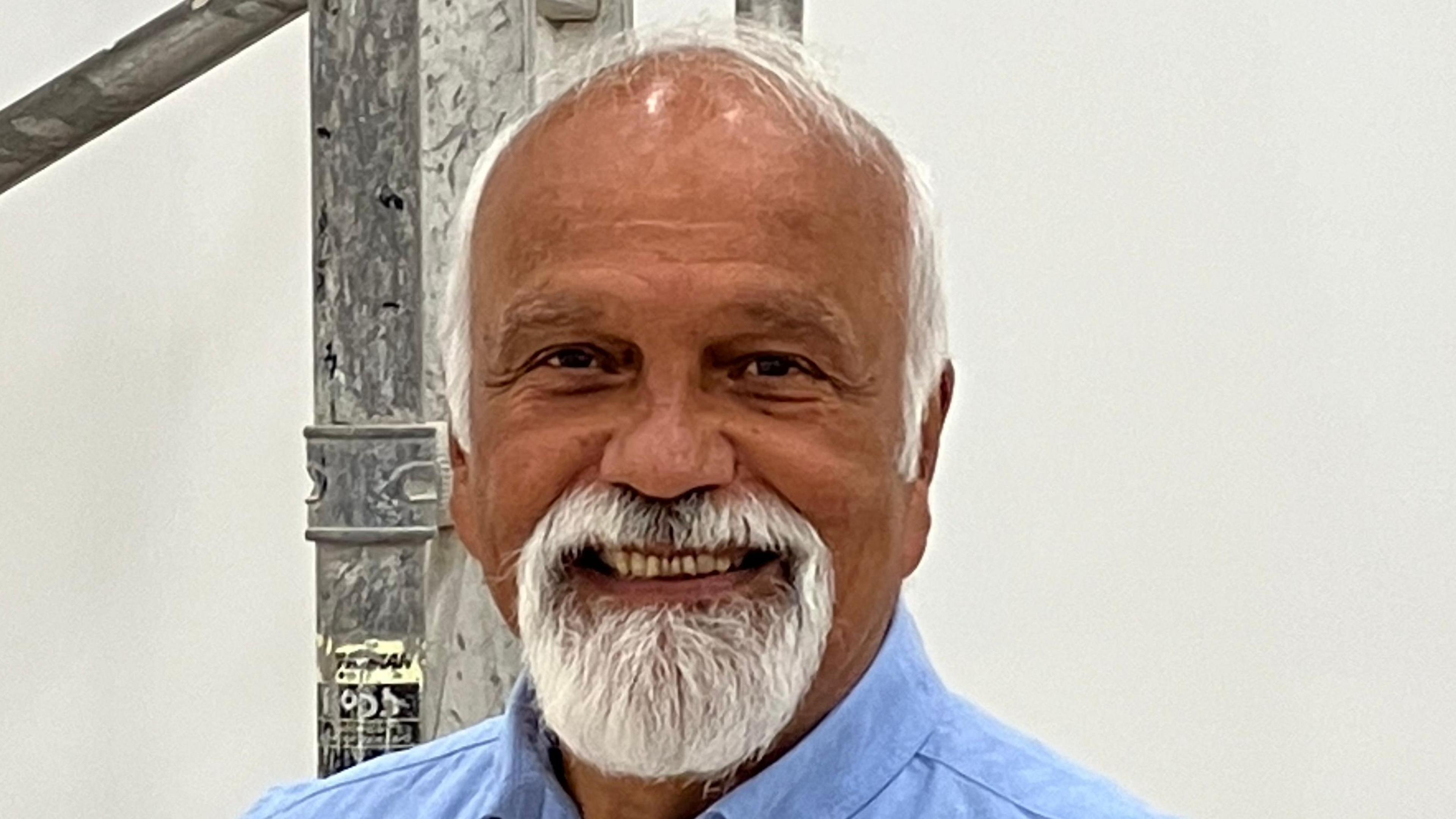 A man with a white goatee beard and a blue shirt standing in front of some scaffolding in a white painted room 