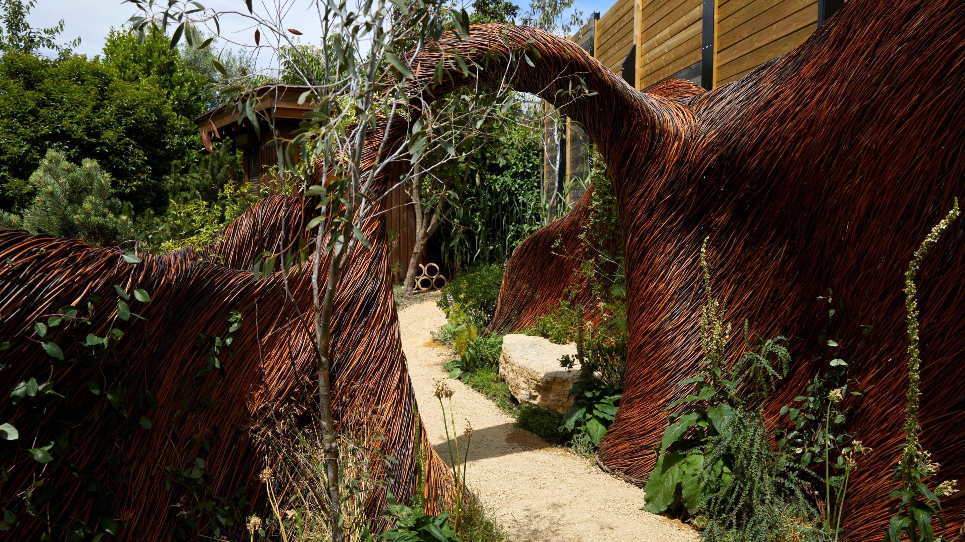 A large wooden, woven arch, with a gravel path beneath it, surrounded by greenery and trees. 