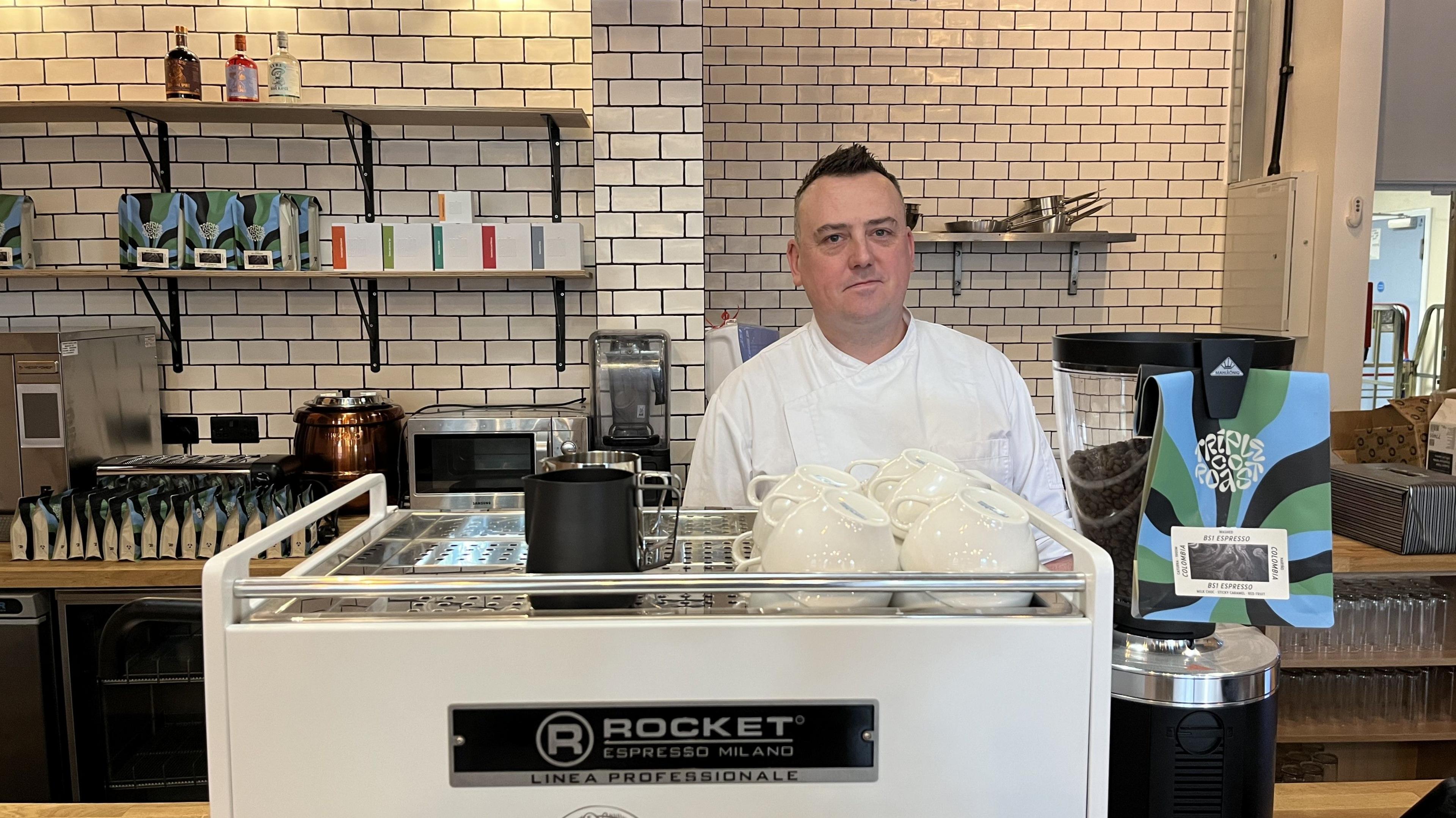 Tom Green wearing a white chef's jacket and standing behind the counter at the Key Cafe. There is a large white coffee machine in front of him. Behind him there is a white tiled wall fitted with wooden shelves, which are stacked with coffee products.