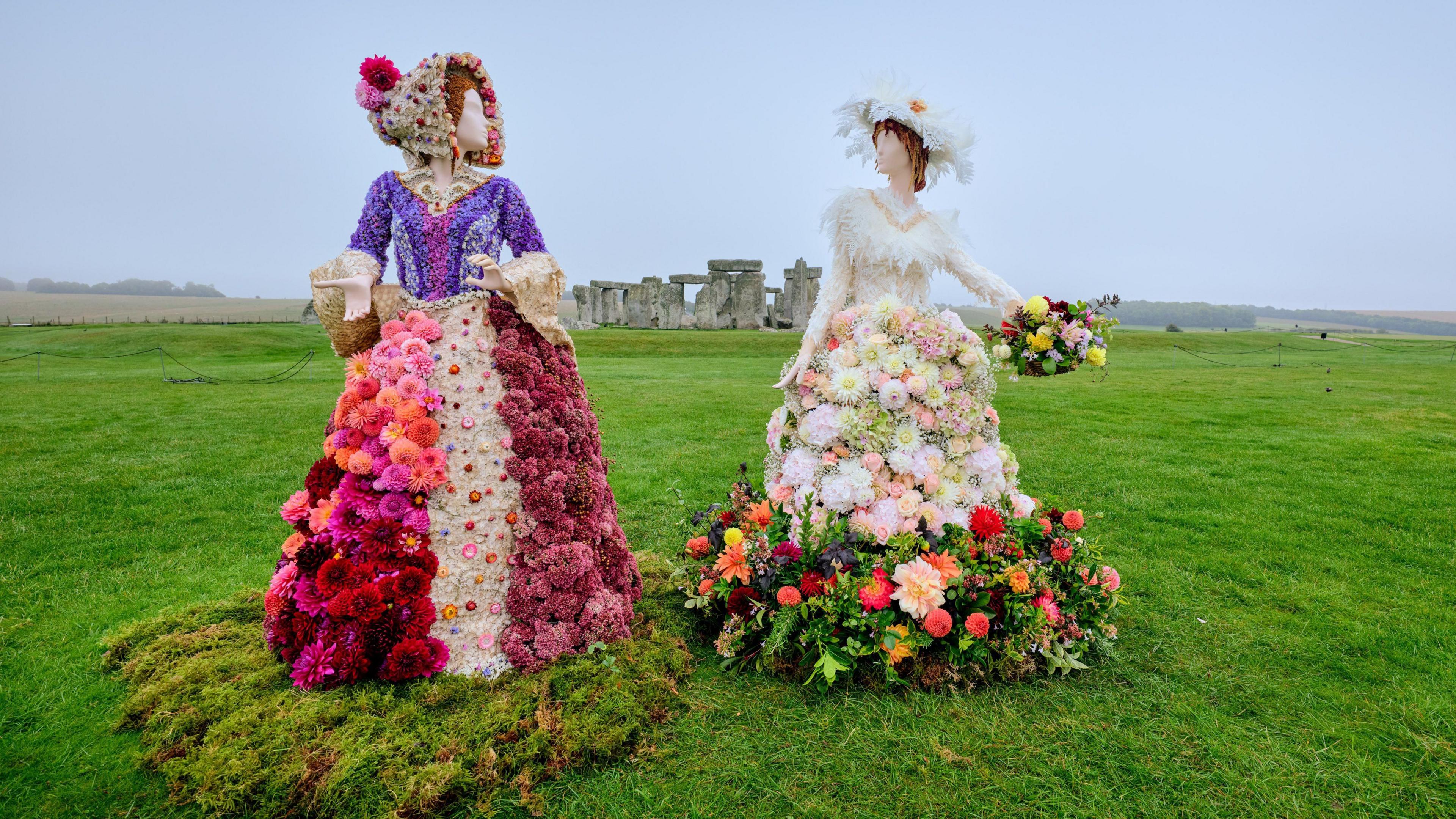 Two Victorian figures made from different colours and types of Dahlia flowers. They are posing elegantly, wearing long dresses, corsets, bonnet hats and carrying woven baskets full of flowers. Behind them is Stonehenge, pictures on a misty and wet day. 