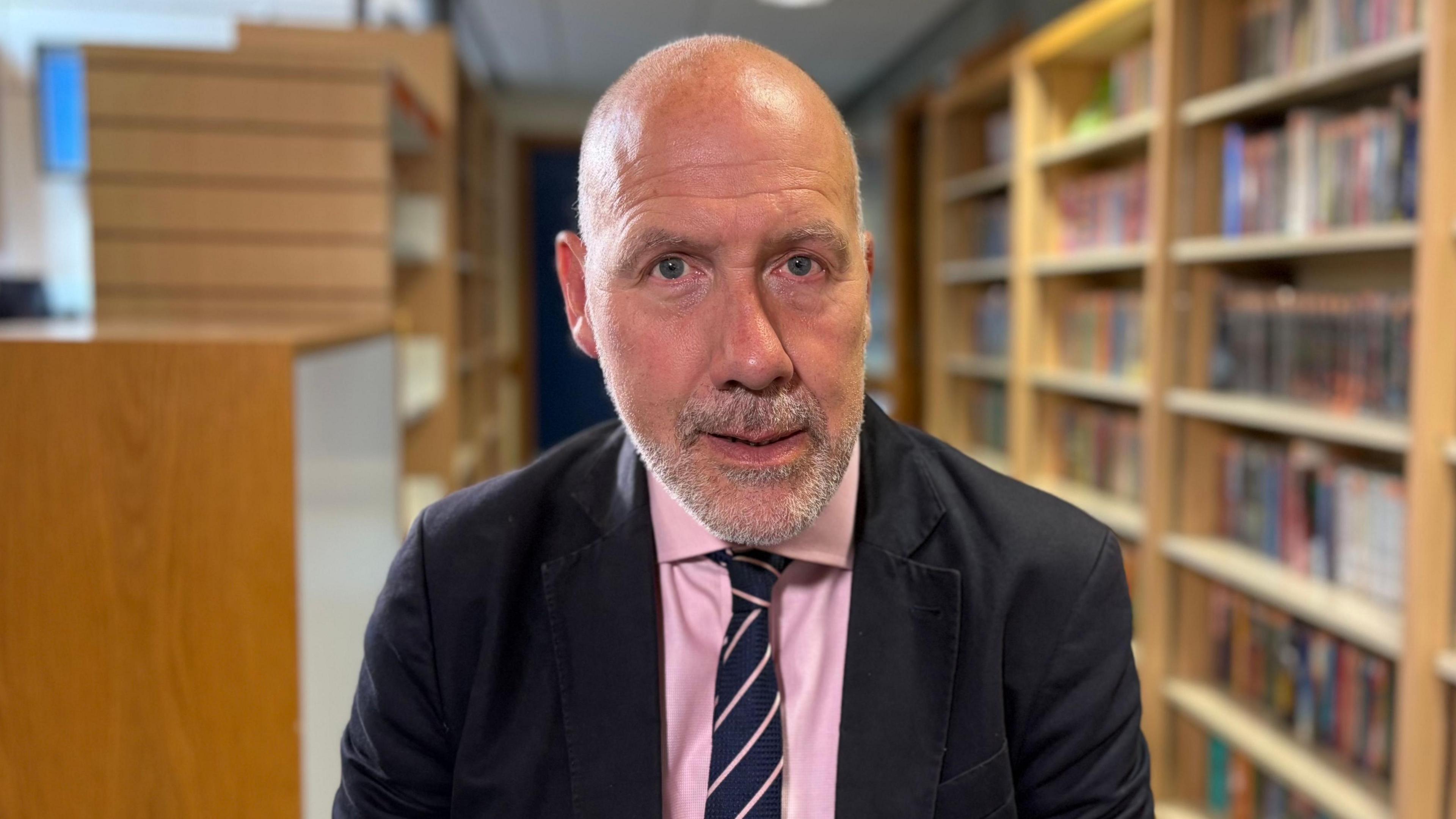 Geoff Barton looking direct to camera. He is sitting down and rows of filled bookshelves are visible behind him, though are out of focus. He is wearing a dark jacket, pink shirt and blue and pink diagonally striped tie. Geoff is mostly bald but has a greying beard. He has blue eyes.