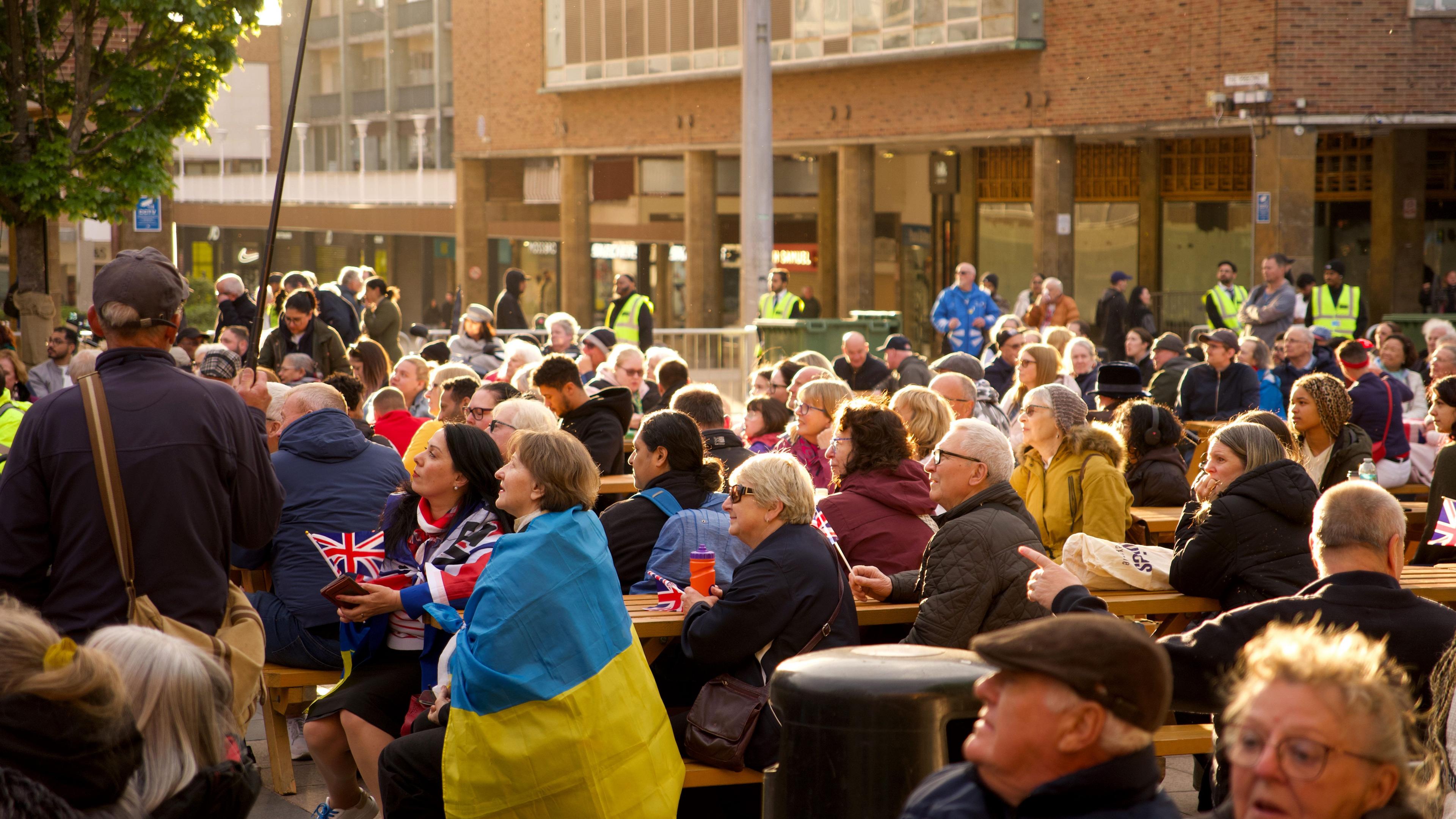 A photo of Coventry's Broadgate square with people sat watching the event on picnic benches. In the foreground, a woman is wearing the blue and yellow flag of Ukraine