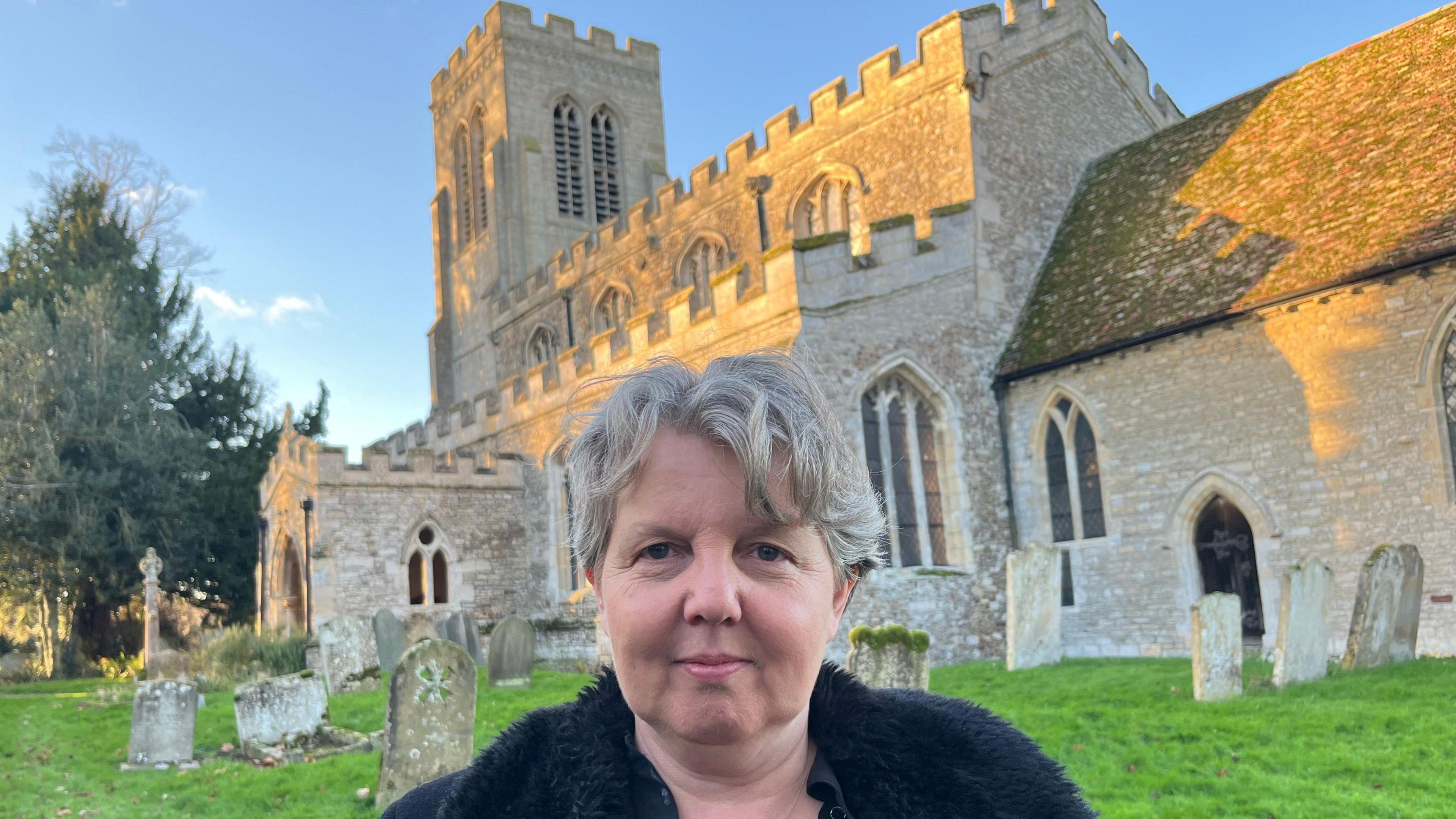 Beverley Vara, the church warden has sandy short hair and a black fur jacket on. She is standing in front of the All Saints Church, which has a tower at one end and there are gravestones in the ground in front of it with trees to the left.