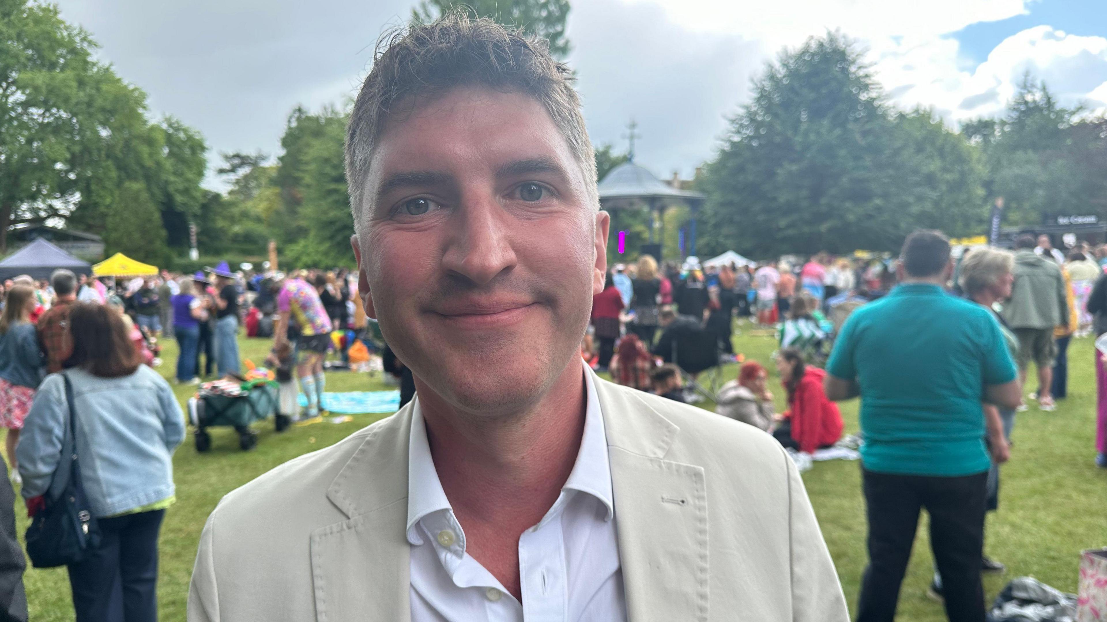 Edward Morello smiles into the camera. He is wearing a cream suit with a white shirt with no tie as he stands in the middle of a busy park of Pride revellers