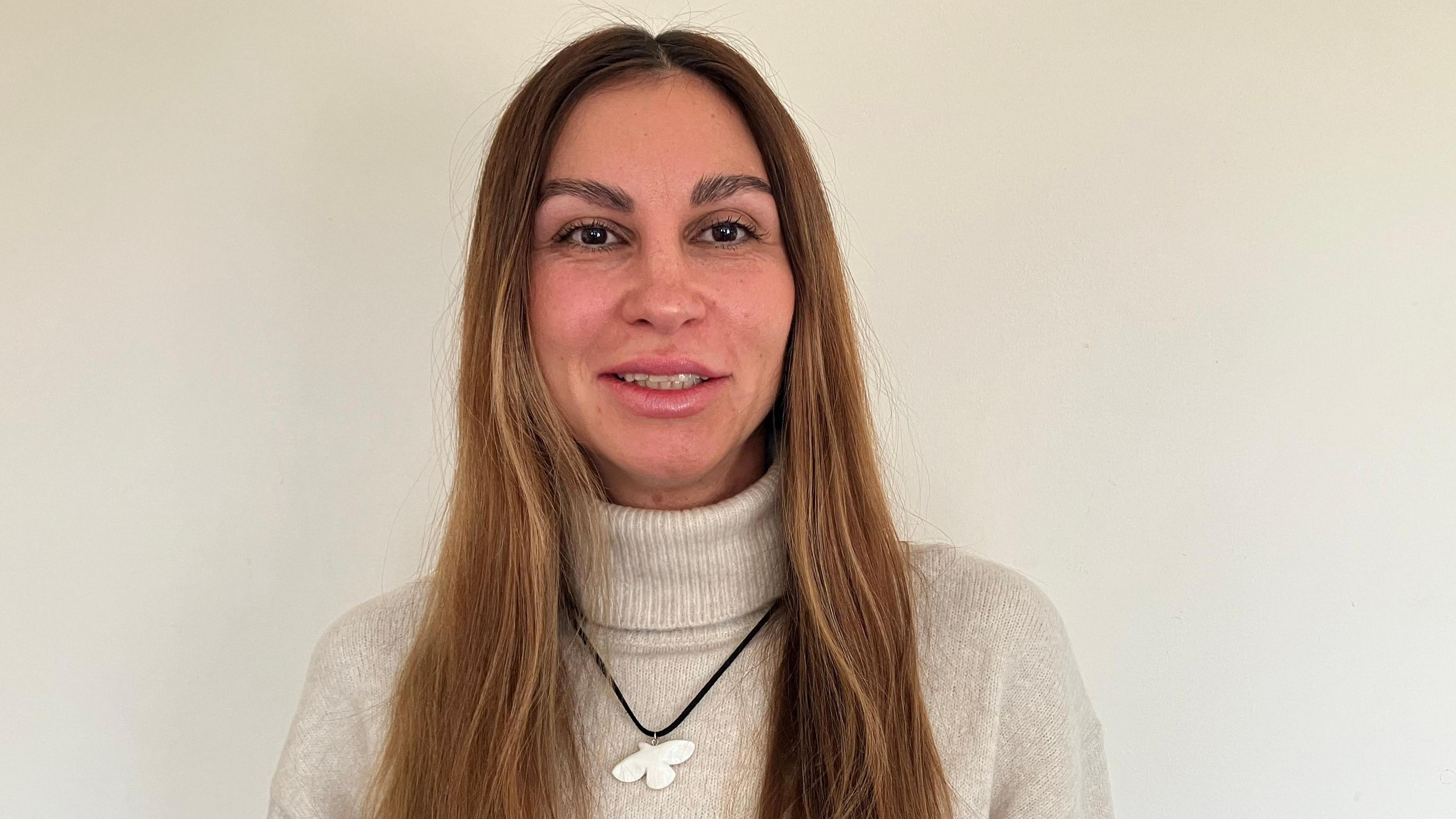 A picture of a woman with longer brownish hair with blonde tints, who is wearing a white roll neck jumper, and black trousers.