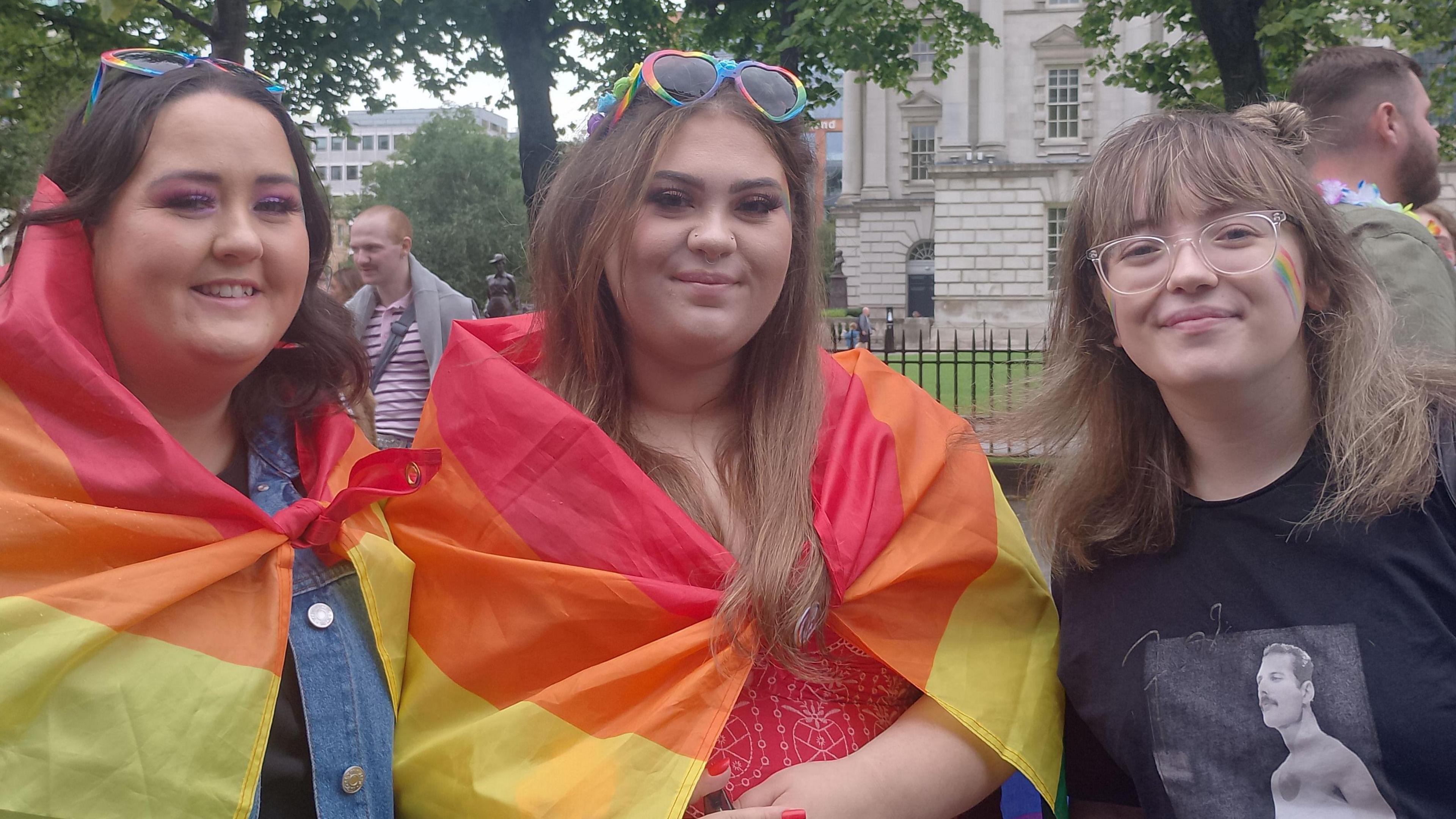 Three spectators at pride. Two have the rainbow flag draped over their shoulders and one has a rainbow flag painted on their face. 