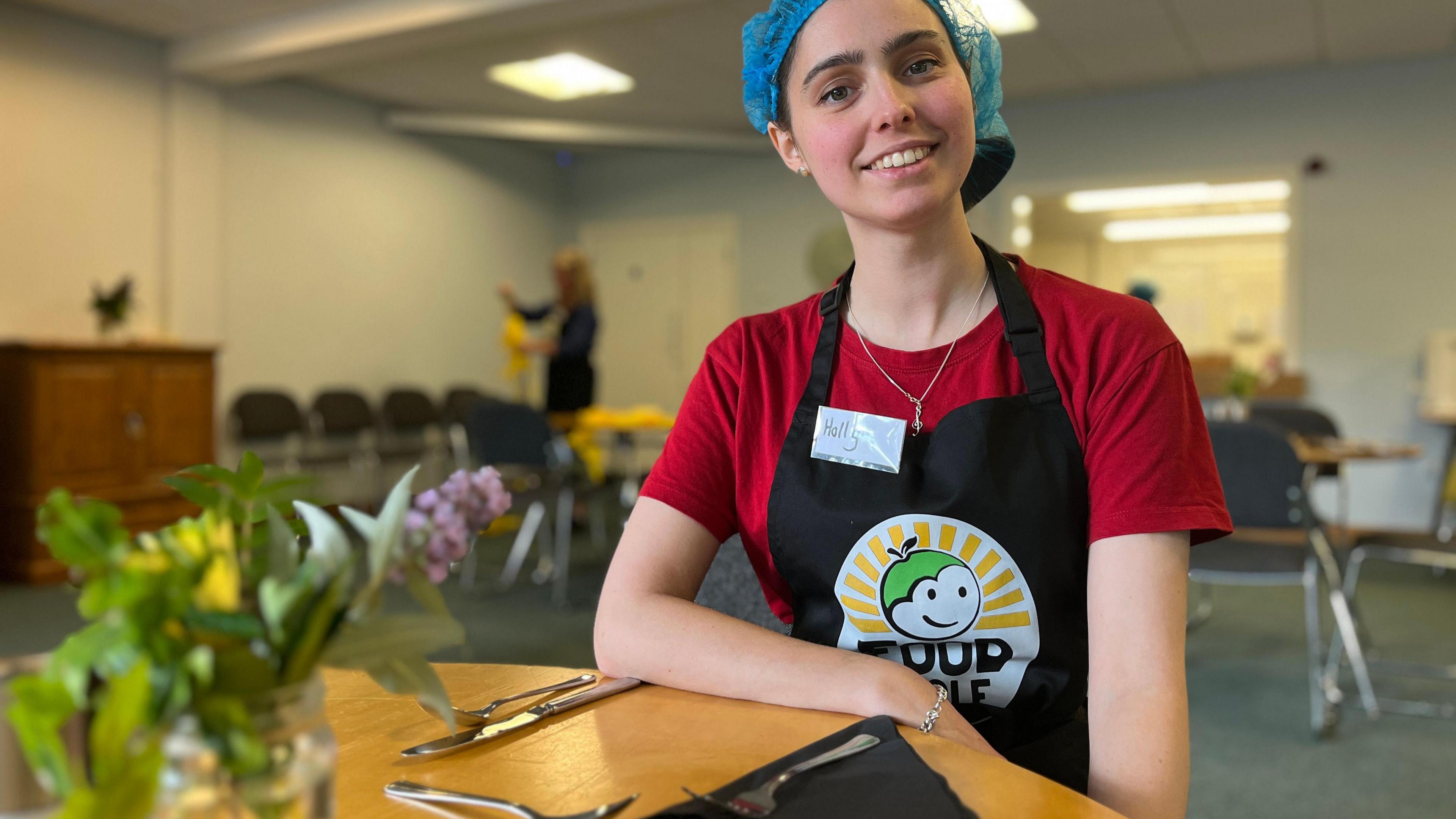 Holly Wright sits at a table in a church hall, with a branded FoodCycle apron and blue hair net