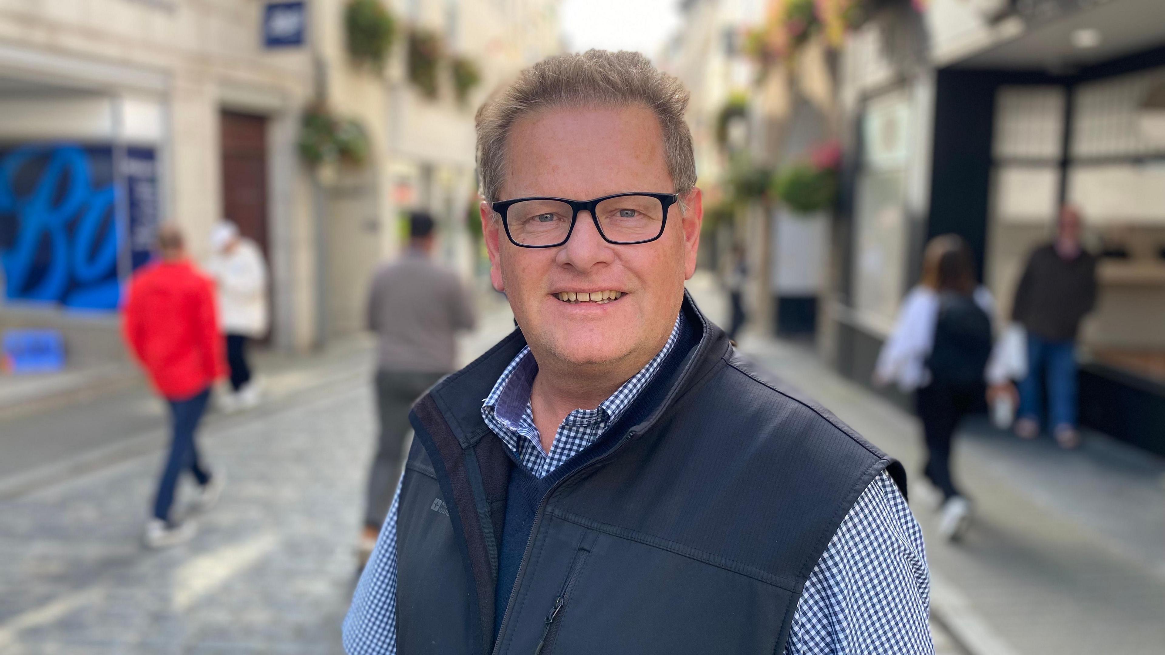 A middle-aged man with salt and pepper hair and glasses is pictured standing on a cobbled street. He is wearing a blue and white checked shirt with a navy bodywarmer over the shirt. There are shoppers in the background.