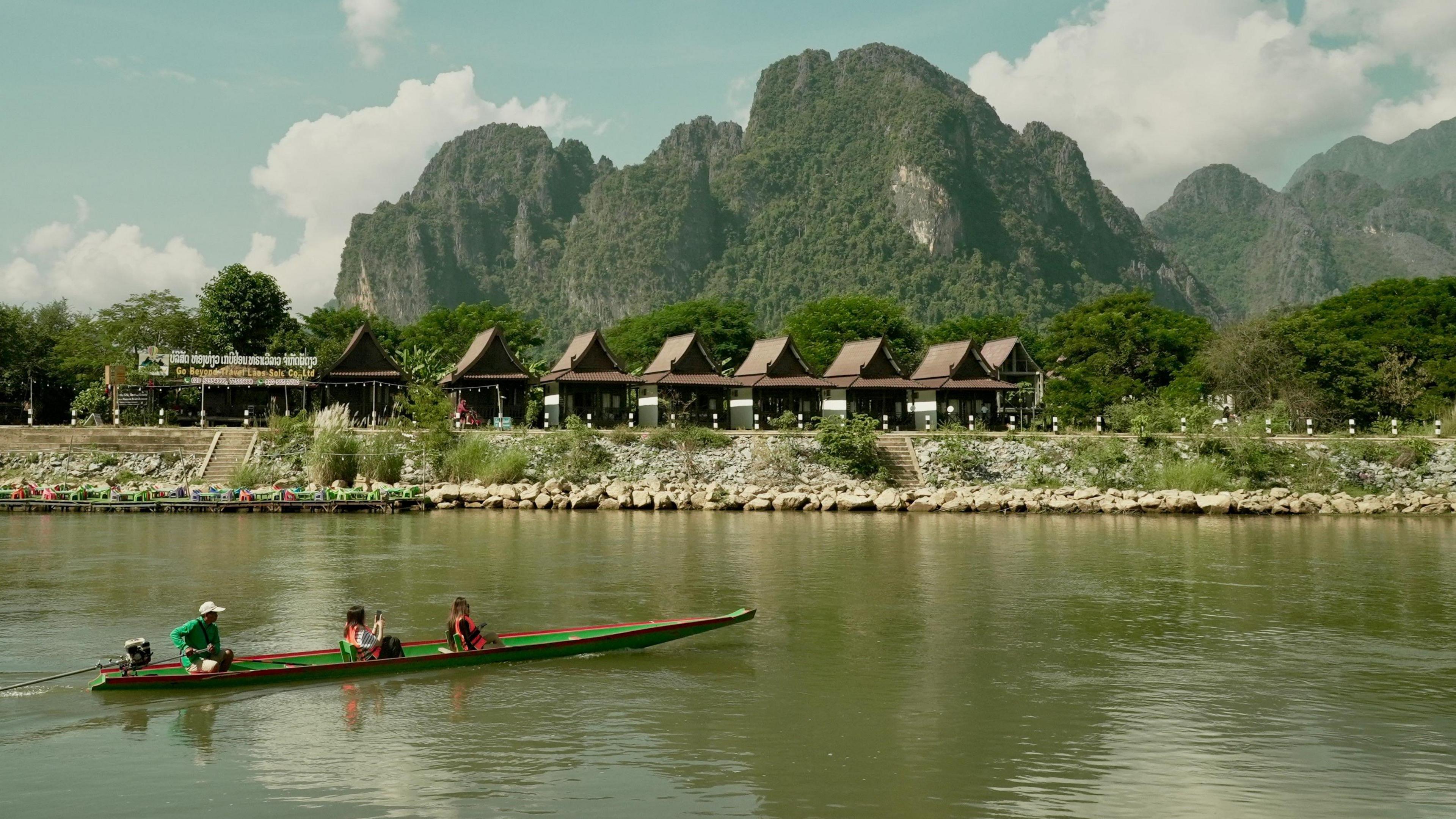 Two tourists sit in a green and red canoe along a river, against a backdrop of jagged wooded peaks, tourist huts and a cloudy blue sky. 