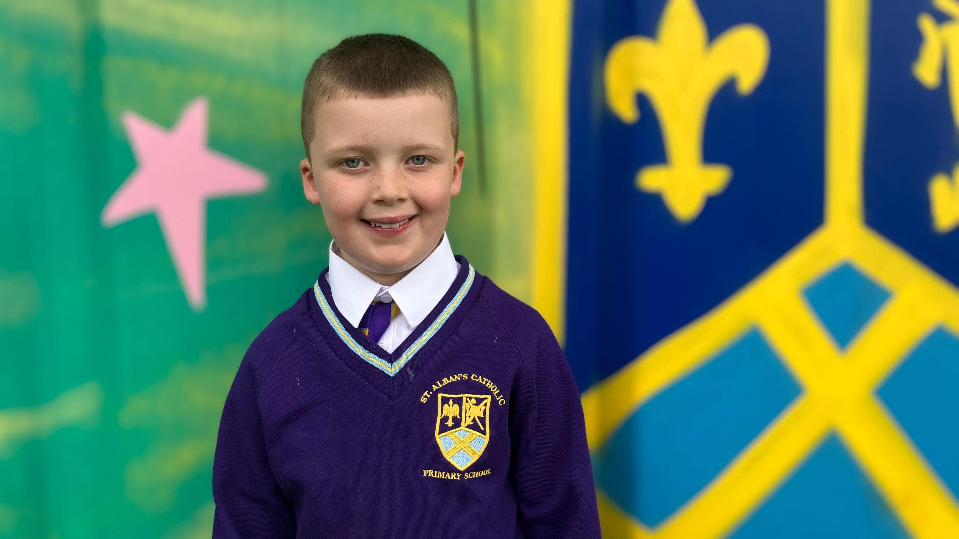 Vinnie, who has short brown hair and wears a purple school uniform, smiles at the camera in front of a painted multi-colour mural.