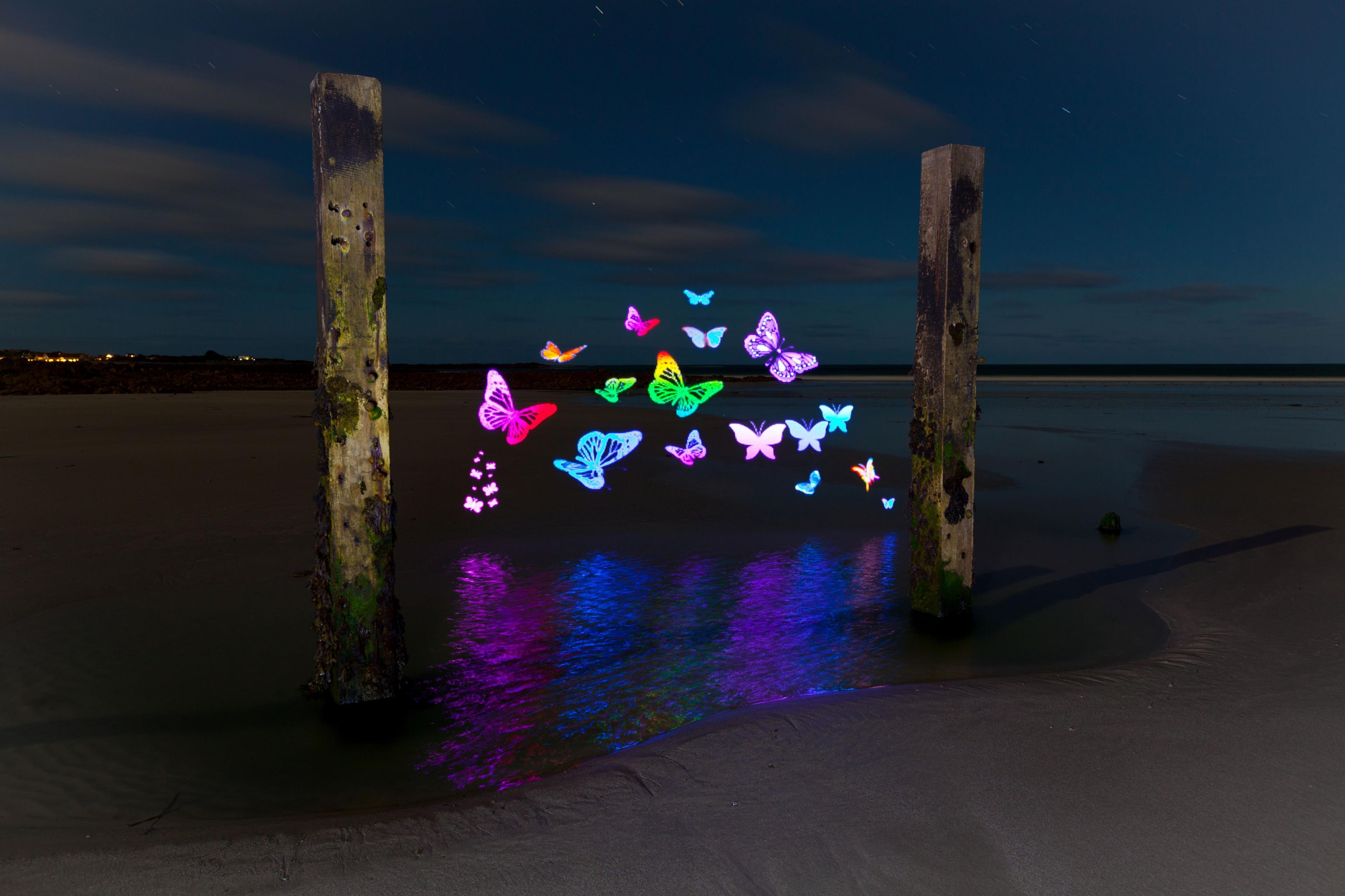 Illuminated butterfly shapes float in mid air above the water in between two wooden posts.