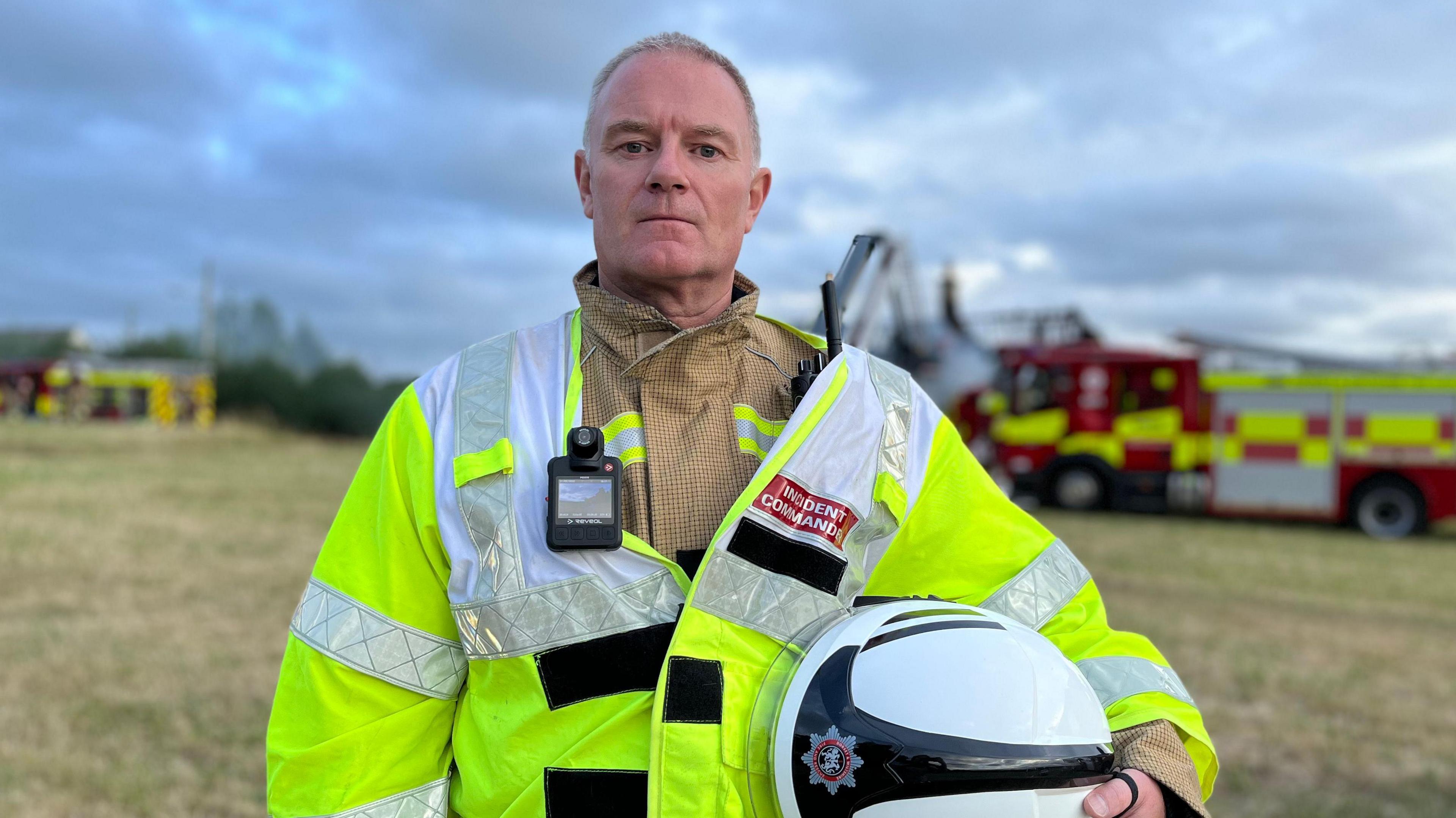 Wayne Jones, shown wearing a high-vis yellow jacket and his firefighting uniform, looks at the camera. He is holding his white helmet with the logo for the firefighting service on it.