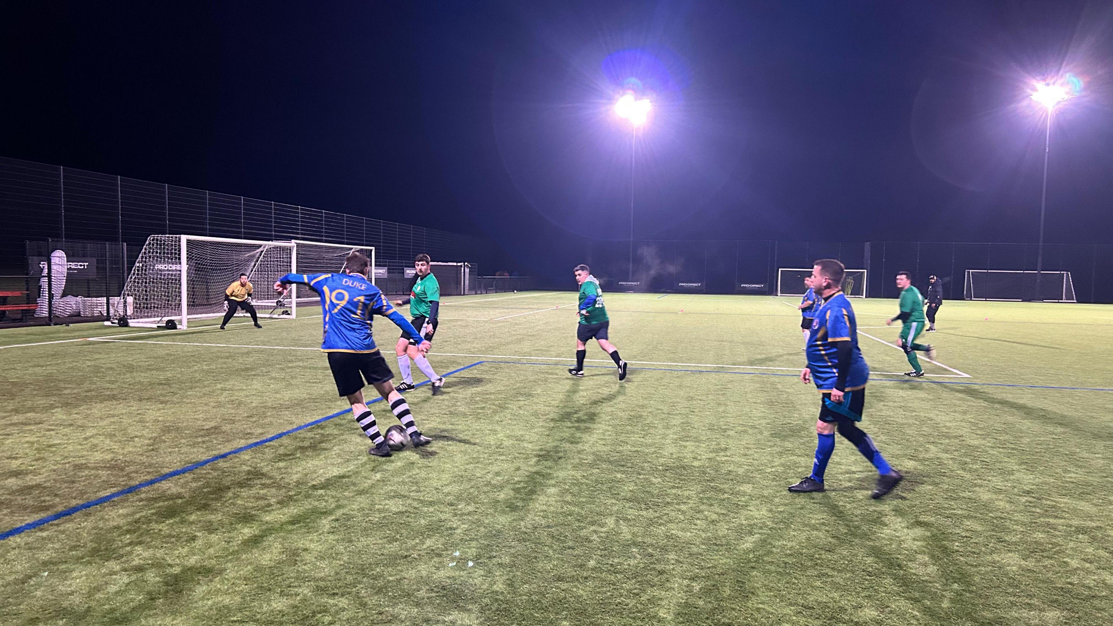 Seven footballers from a Man v Fat session in Exeter playing on one of the pitches at Exeter City FC's Cliff Hill training ground. Three of the players are in blue, three are in green and the goalkeeper is in yellow. One of the players, with 91 on his back, is dribbling with the ball towards the goal. 