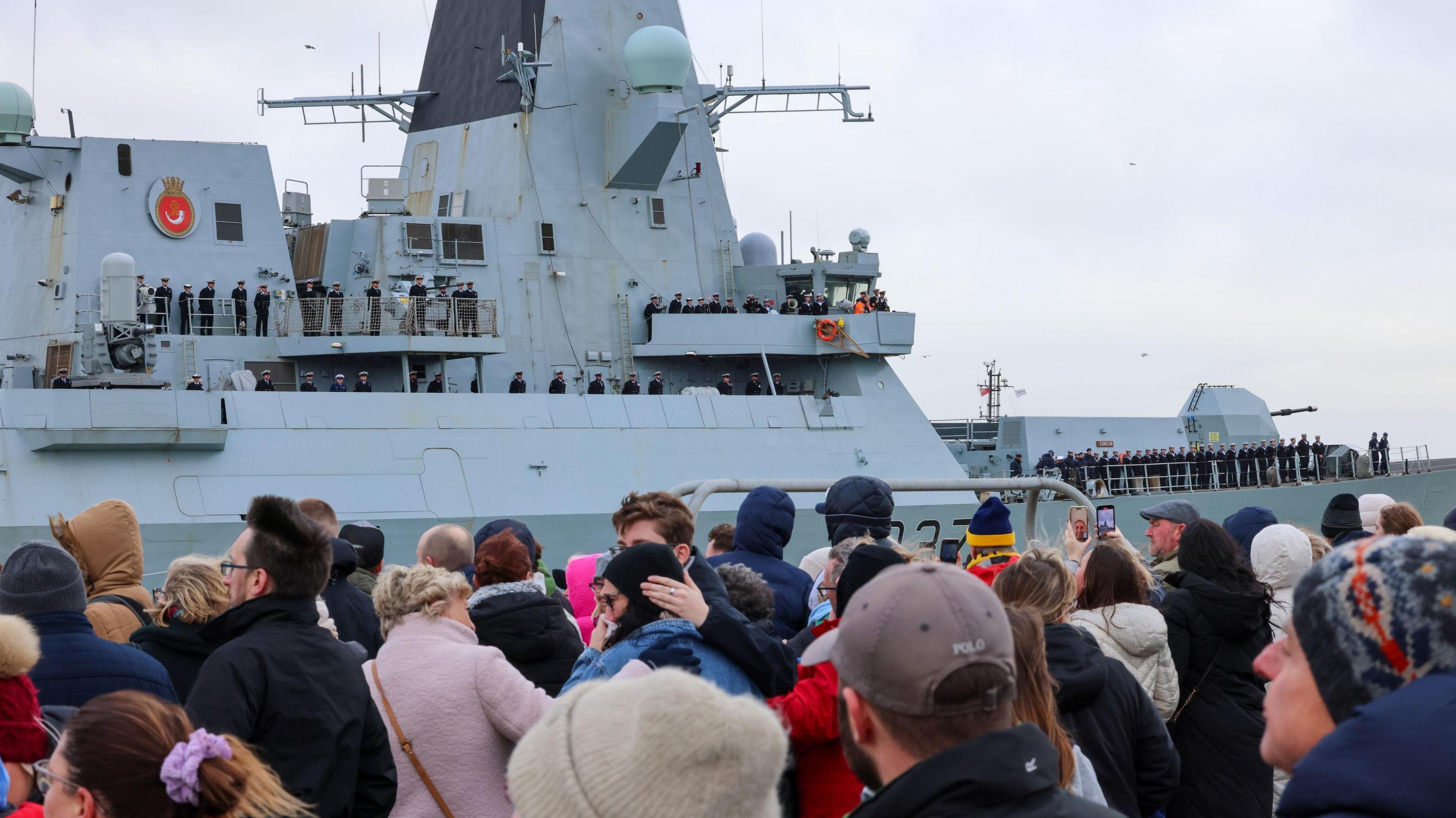 Crowds of people are lined up along Portsmouth harbour, wearing warm clothing and watching as HMS Duncan enters the harbour. The destroyer is a large pale grey coloured ship and sailors in uniform can be seen lined up along the ship's deck.