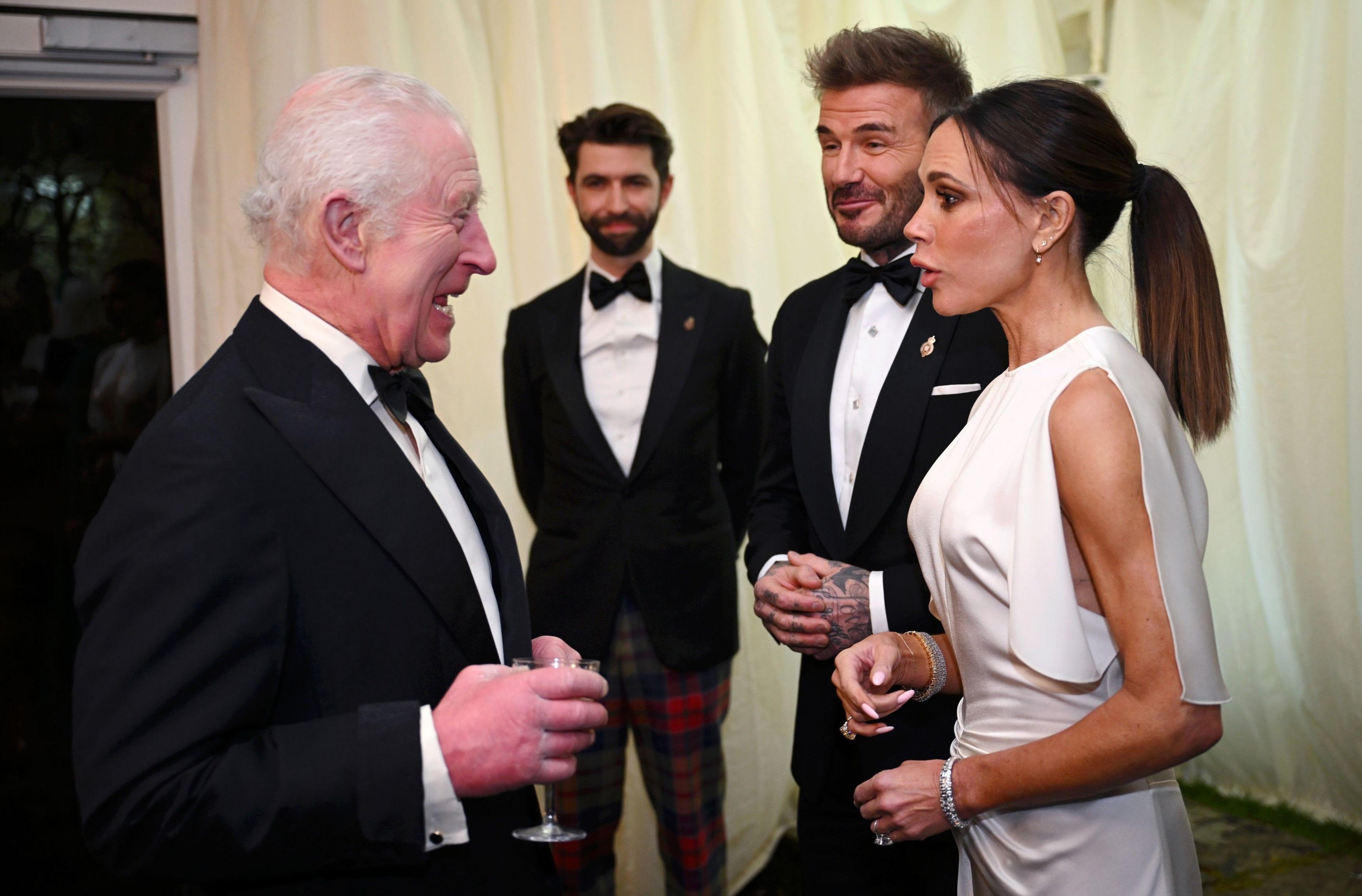 King Charles, wearing a black tie outfit at a formal event, talks to Victoria and David Beckham. Victoria is wearing an all-white formal dress and David is also wearing a tuxedo and bow tie