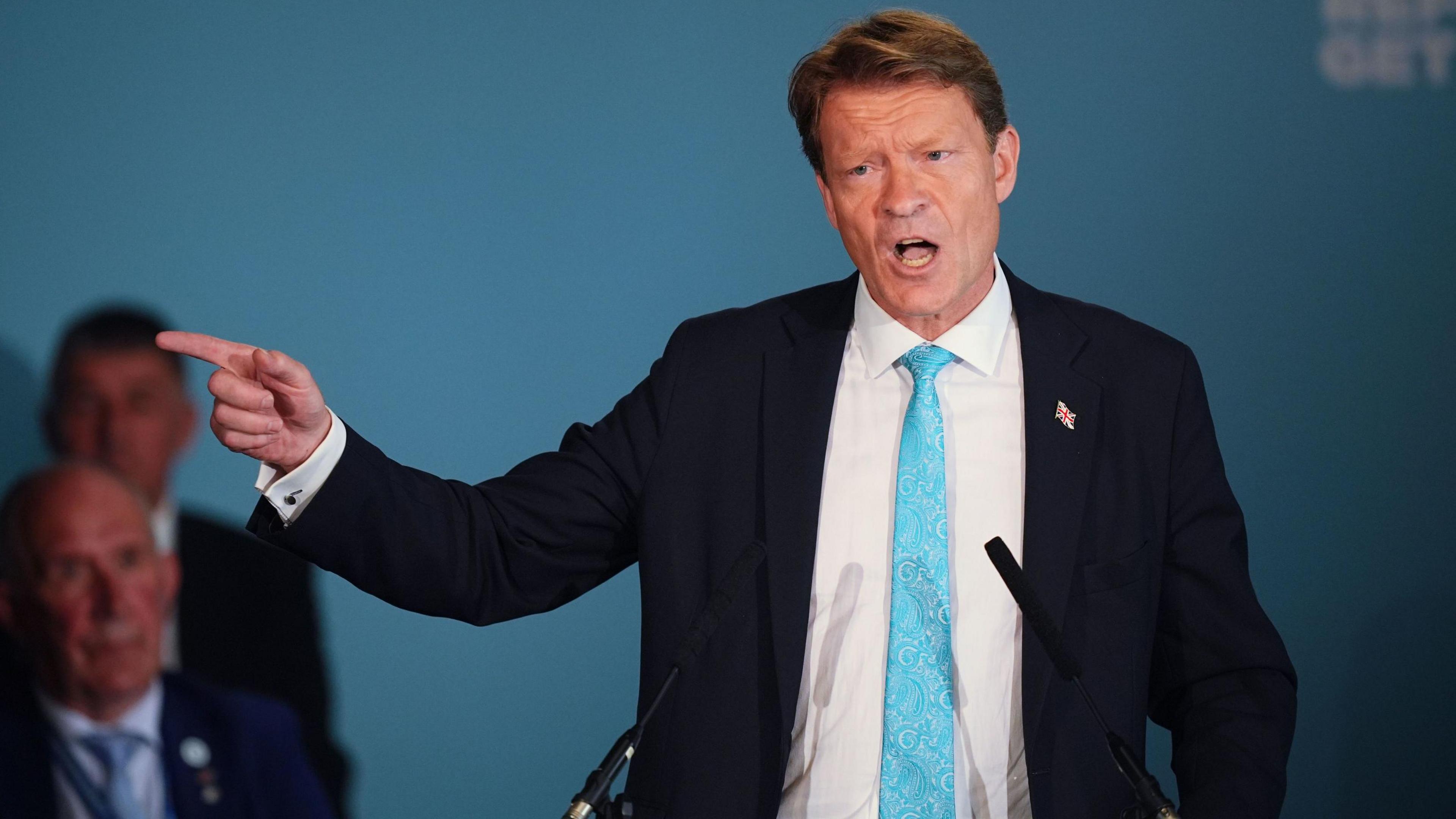 Reform UK deputy leader Richard Tice speaks during a press conference at the Royal Horseguards Hotel in London. He is pointing with his finger to the left. He is wearing a black suit jacket, white shirt and a blue tie. He has short brown hair.