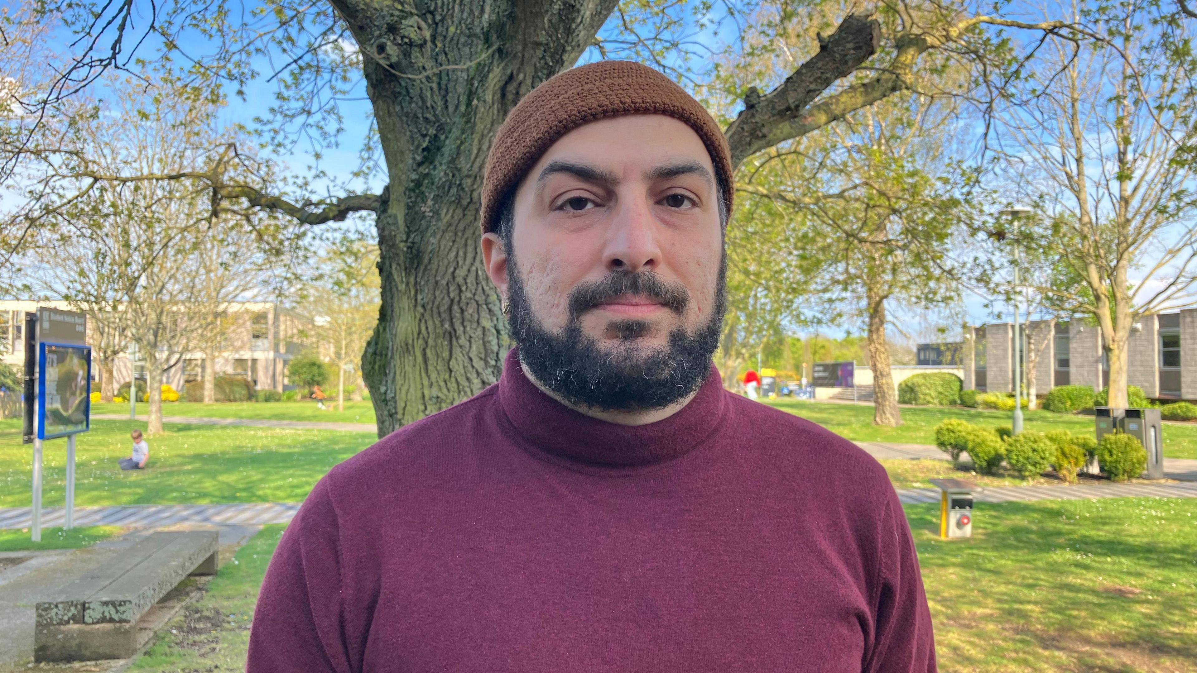 Dr Taymaz Azimi, wearing a burgundy rollneck jumper and a brown knitted hat. He is standing in front of a tree on the grounds of the University of Kent. 