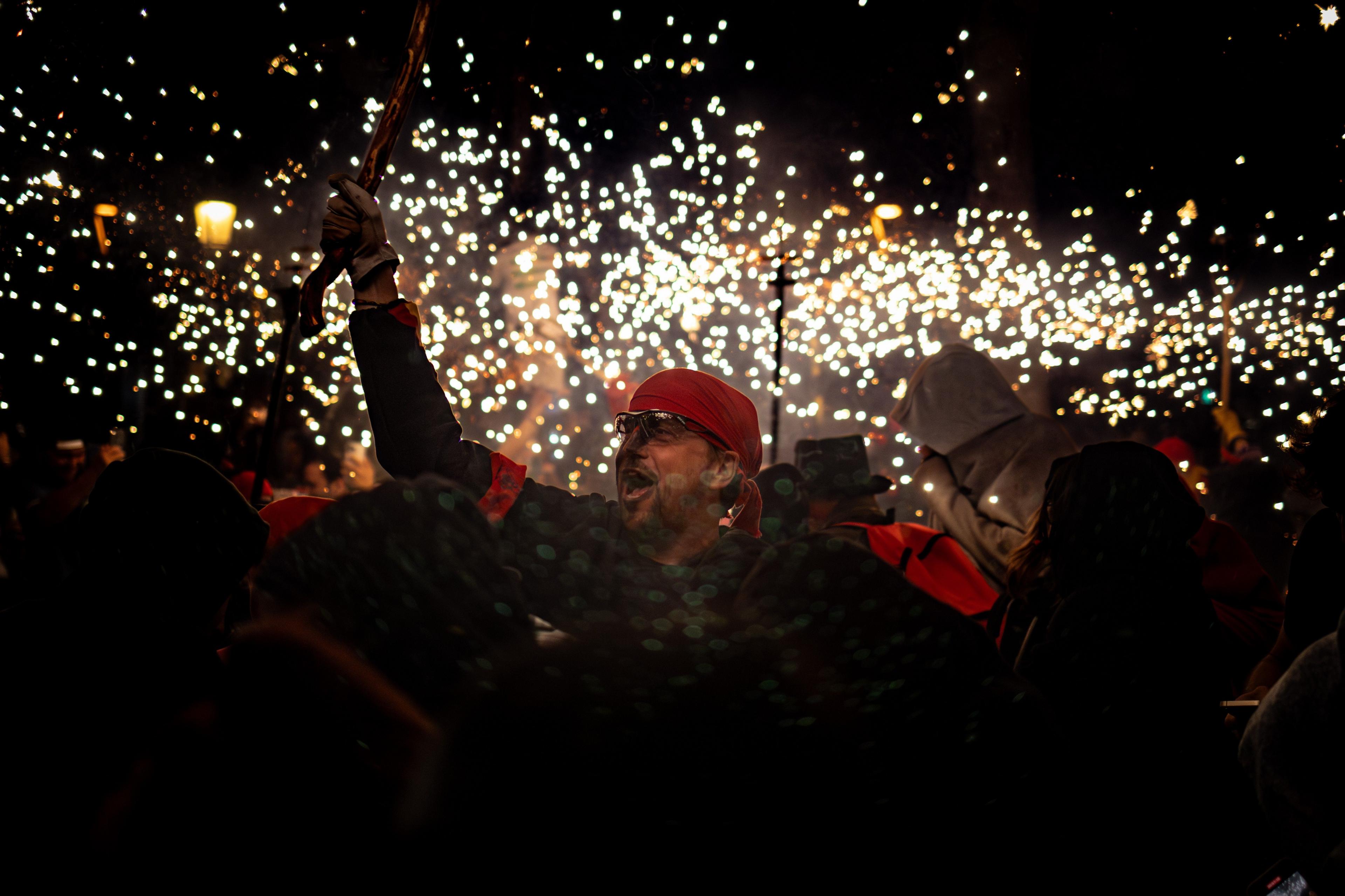 A man in a red bandana smiles and holds his arm aloft. He is part of a crowd of people celebrating and there are flying sparks in the night sky.