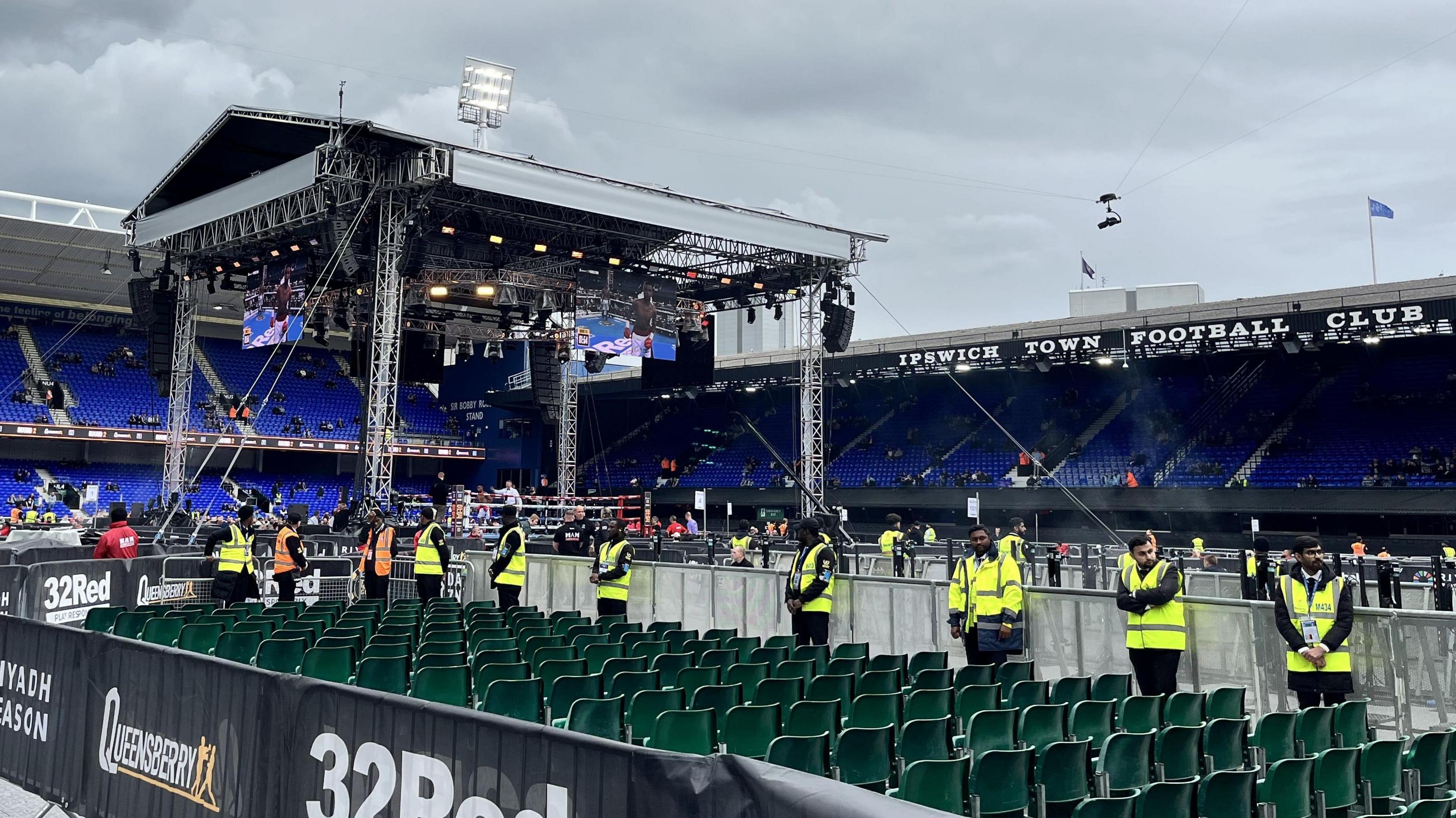 View from the left side of the boxing arena from behind rows of chairs. Security in yellow hi-vis jackets are lined-up outside of the barriers next to the boxing ring.