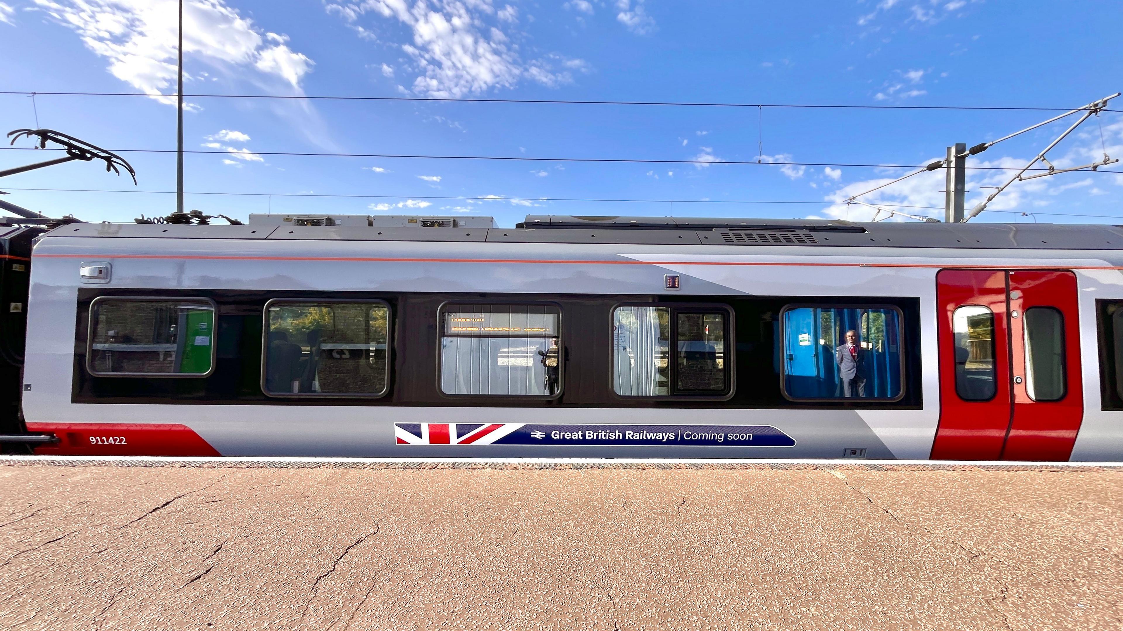 A wide shot of a train carriage standing at a platform. It is a bright day, with a blue sky visible above the train and only a couple of wispy clouds. A smartly dressed looking member of staff can be seen reflected in one of the carriage's windows. Above the train are power lines. Along the bottom of the carriage is a logo, which includes a small portion of the red, white and blue stripes of the Union Flag, the parallel lines of the railways symbol and the text 'Great British Railways - Coming soon'.