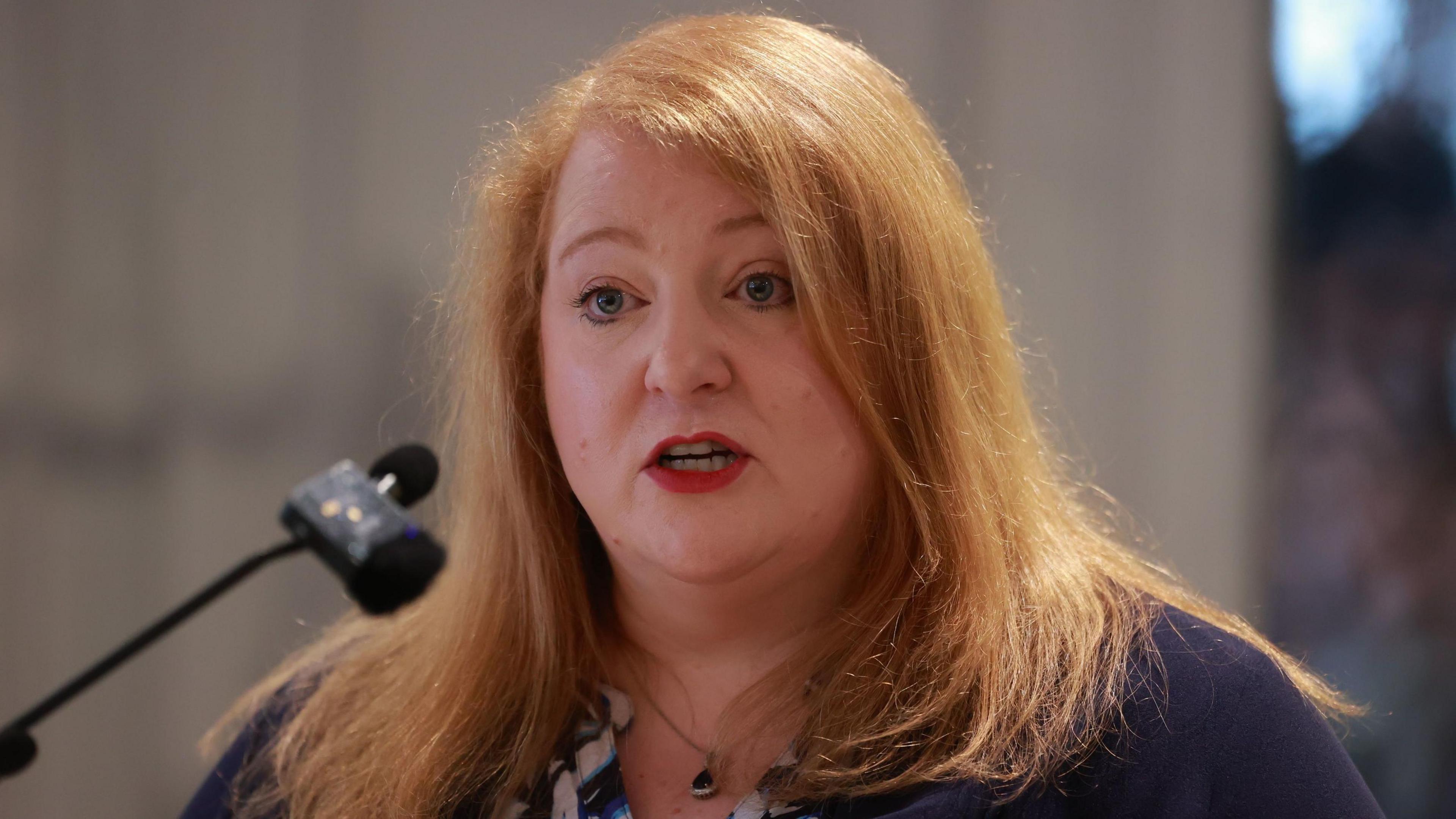 Naomi Long - a woman with shoulder-length, ginger hair is speaking into a small microphone. She is wearing a silver necklace with a black gem, a blue and white patterned top and a navy blue jacket.