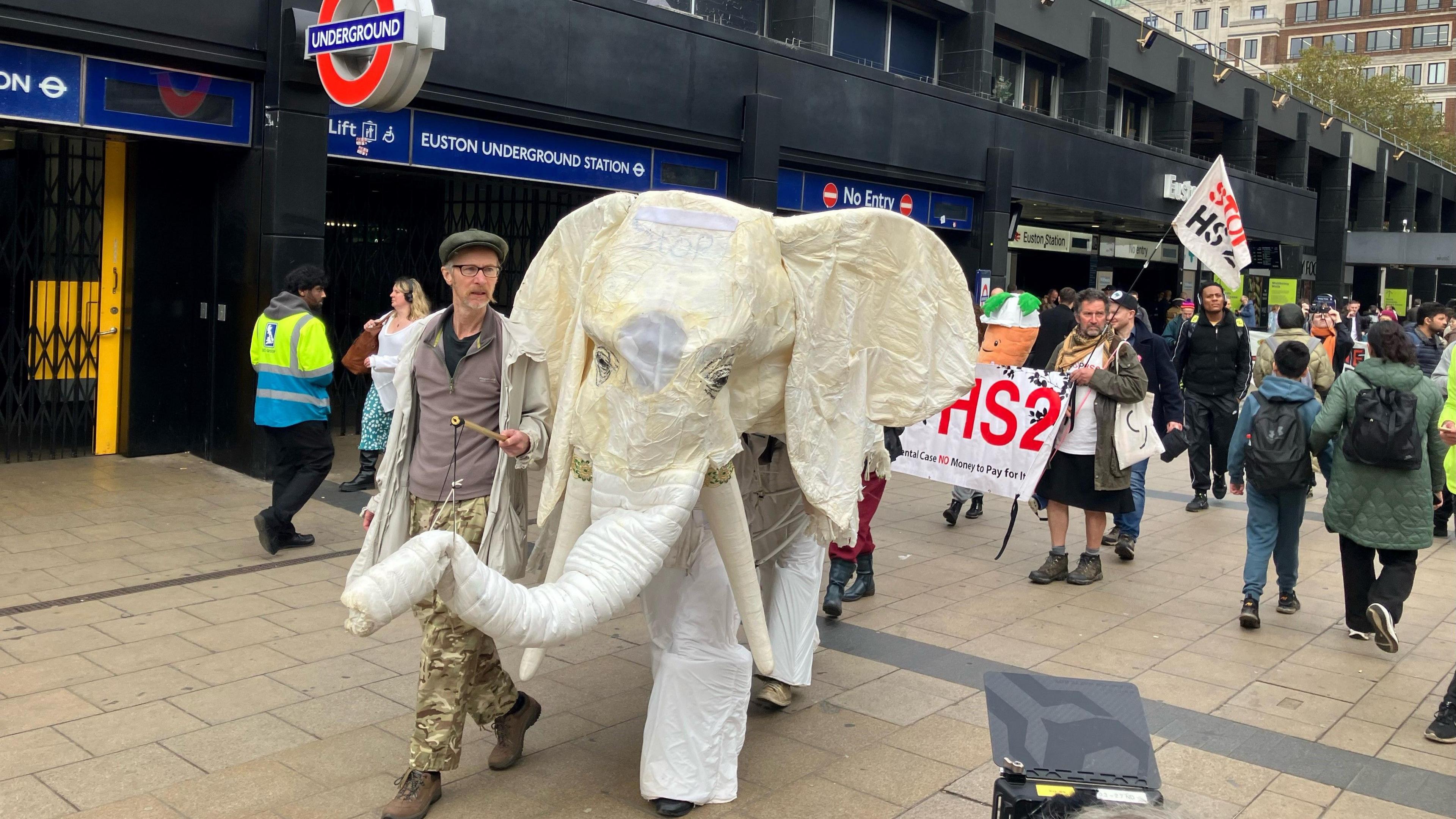 HS2 protesters outside Euston Station in London, led by a person with a large, white model elephant comprising of two people. Signs reading "Stop HS2" are visible.