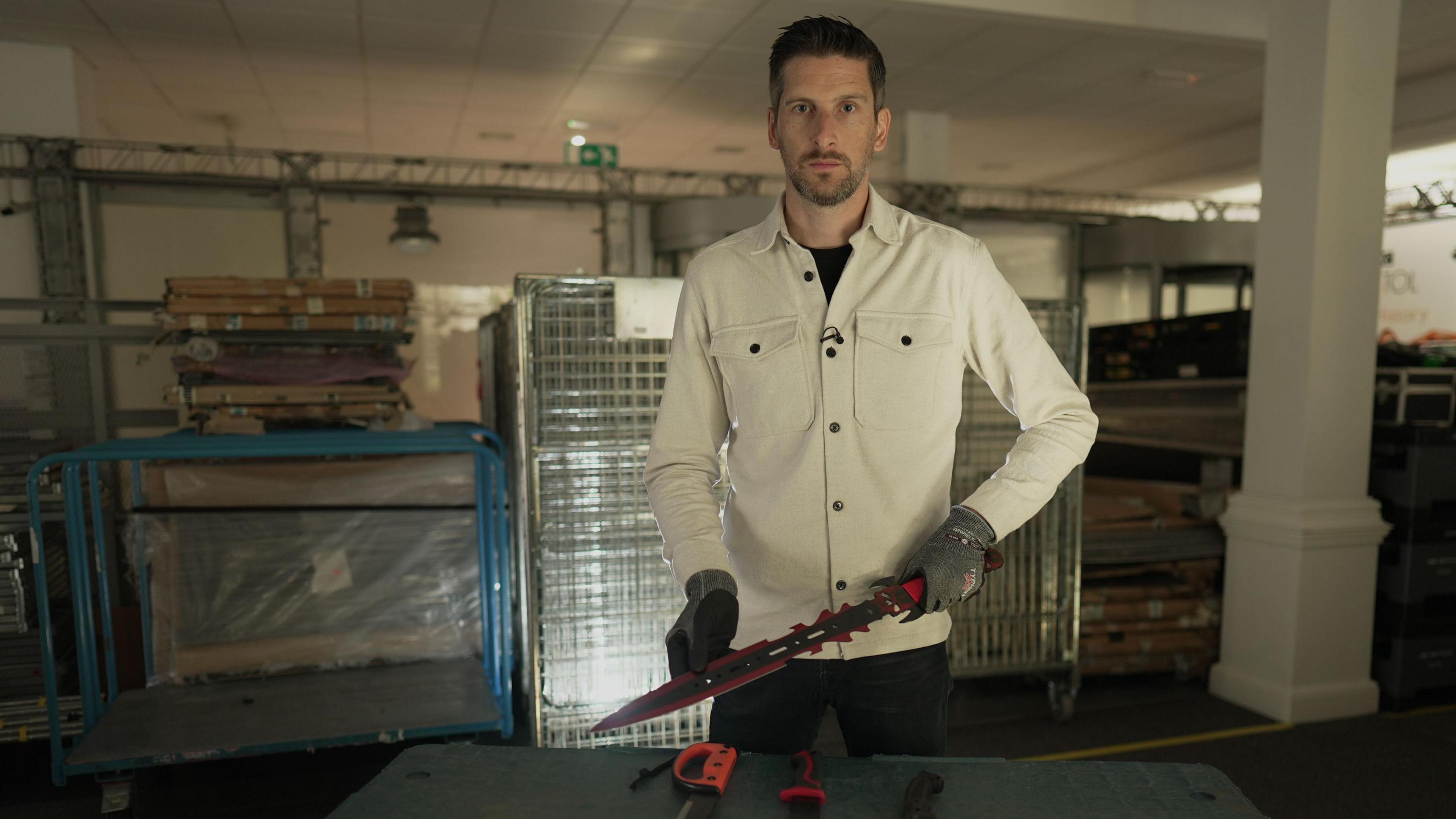 BBC News's Dan Johnson holding one of the purchased knives. He is holding the large knife in two hands while wearing gloves, looking directly to camera