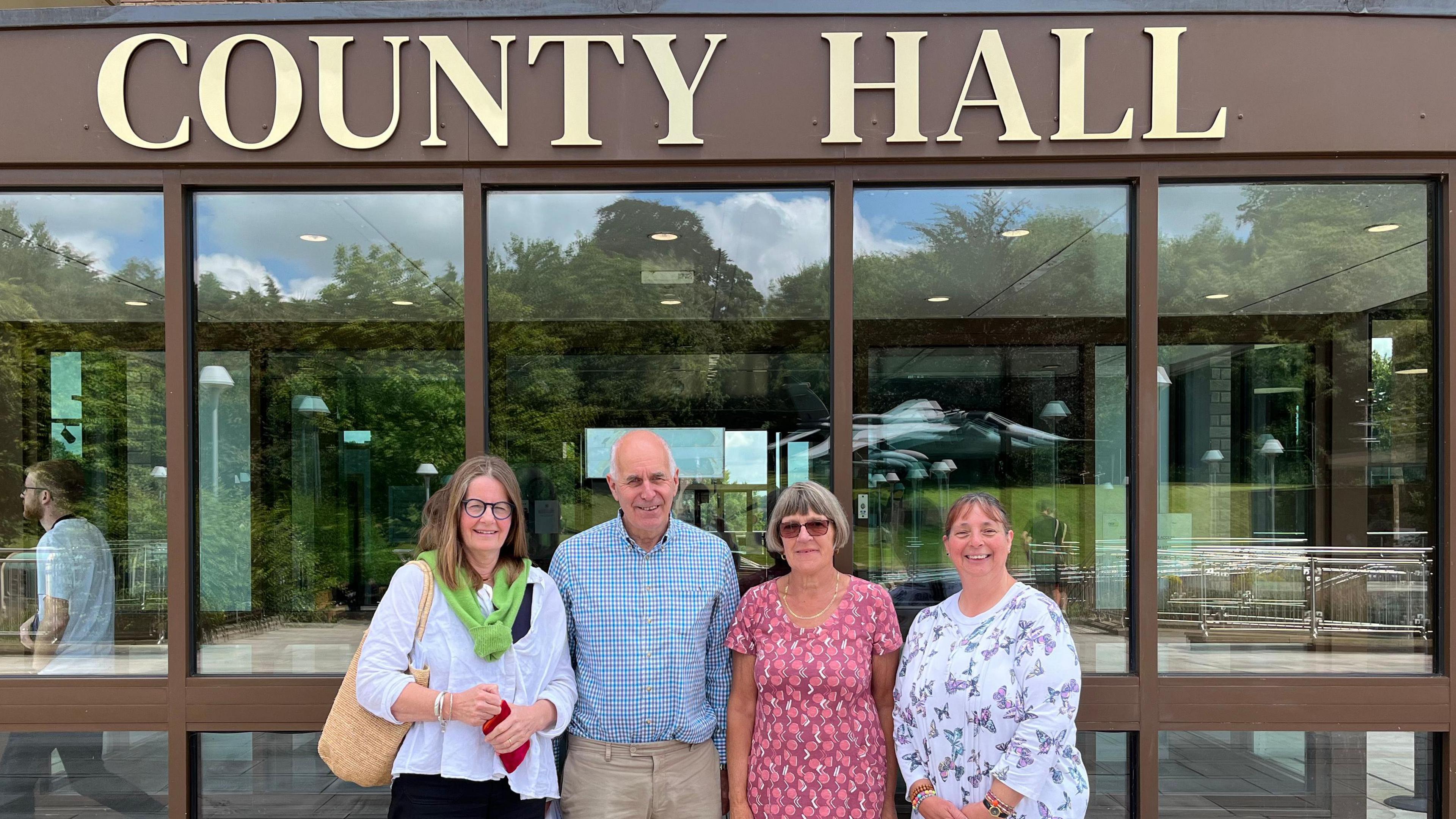 Four people standing outside the Norfolk County Council building. From left to right a woman with black glasses and a white shirt, next to her a man in a blue checked shirt, next to him a woman in a pink patterned dress and sunglasses and on the end is a woman  wearing a white top with butterflies on it