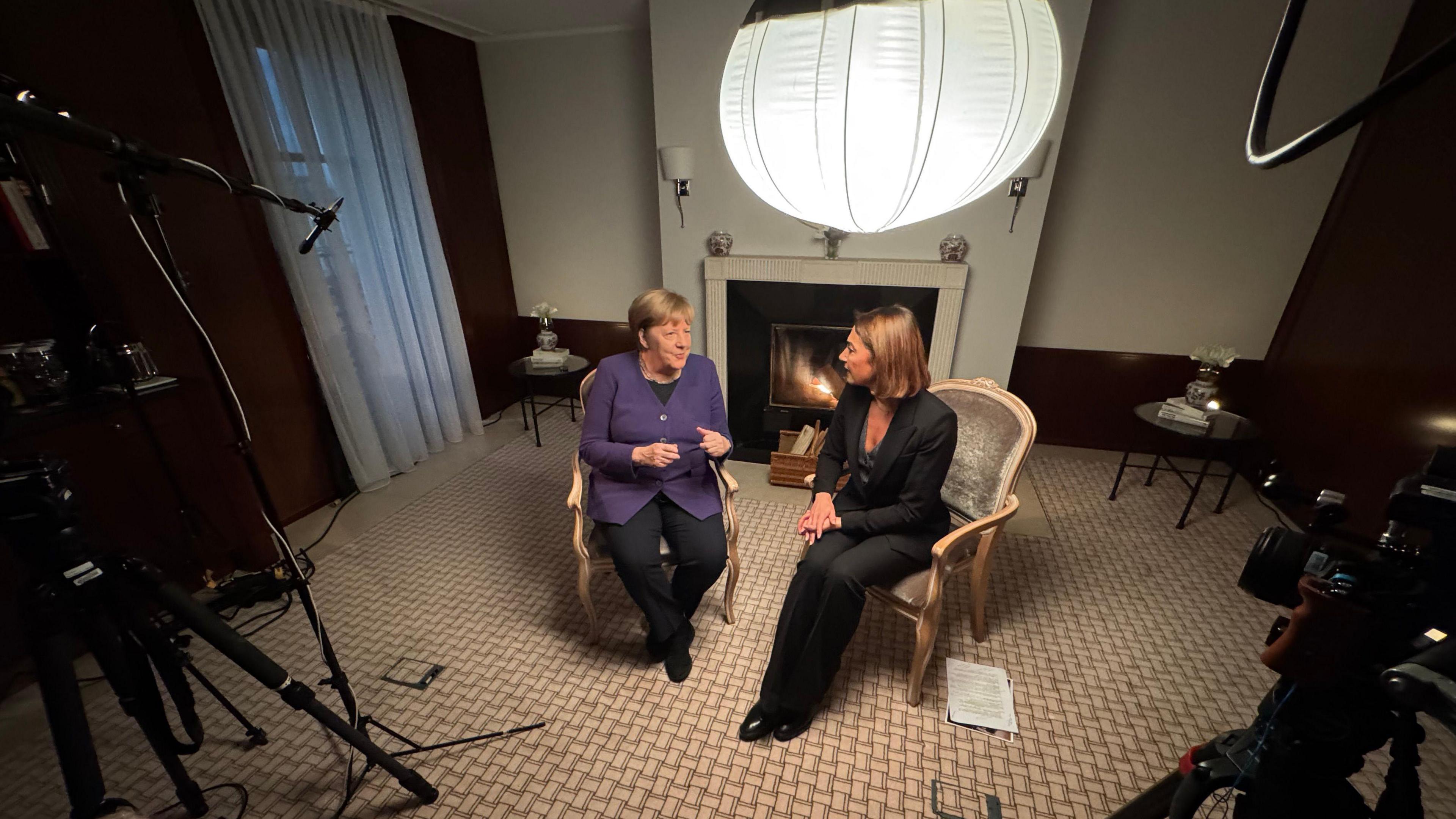 Former German Chancellor Angela Merkel sits talking about her time in office with the BBC's Katya Adler in a hotel room. Their chairs are facing each other and there is a variety of broadcast equipment in view