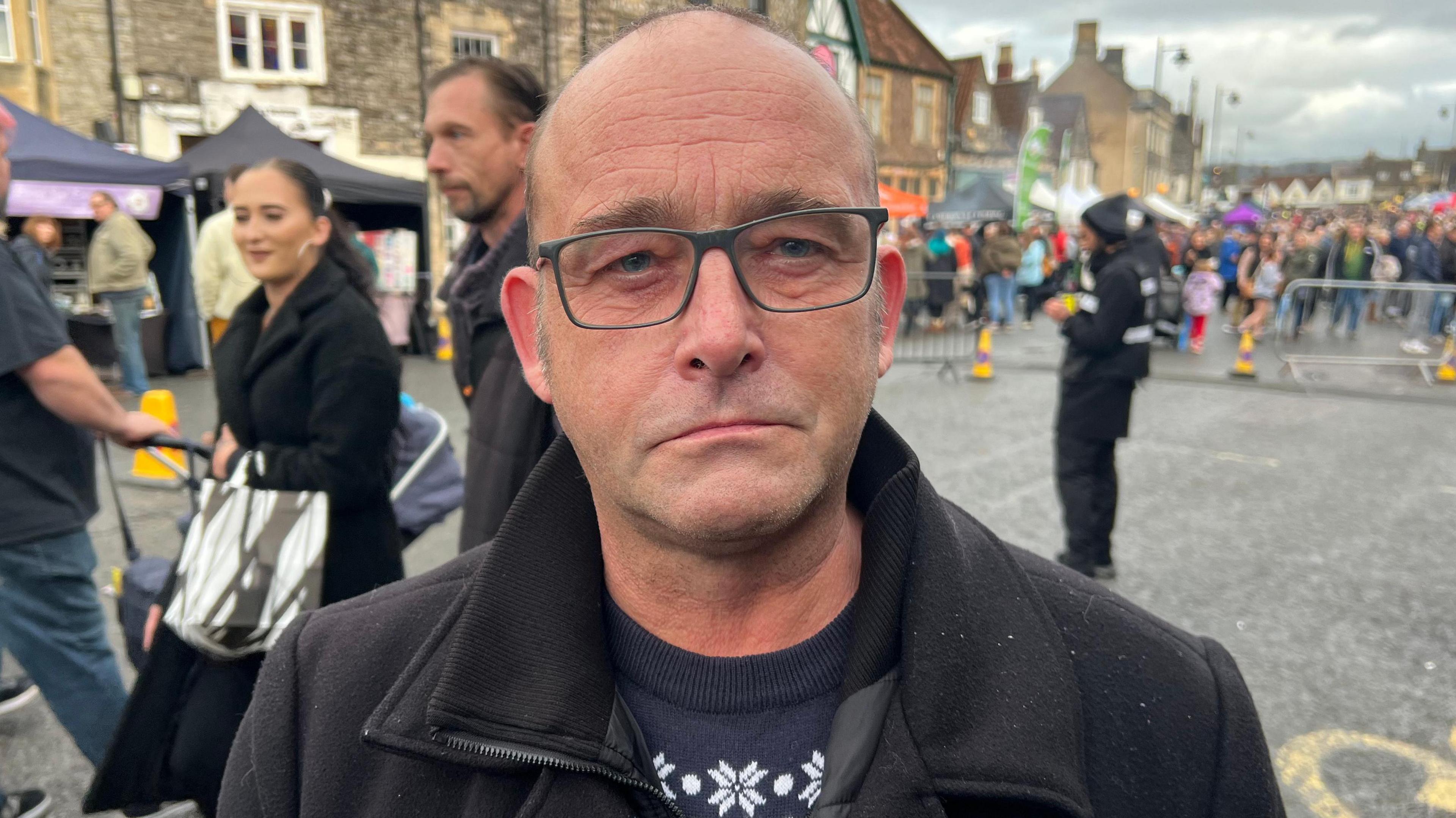 A man wearing glasses a dark Christmas jumper and dark coat standing in front of a crowd of people looking into the camera 