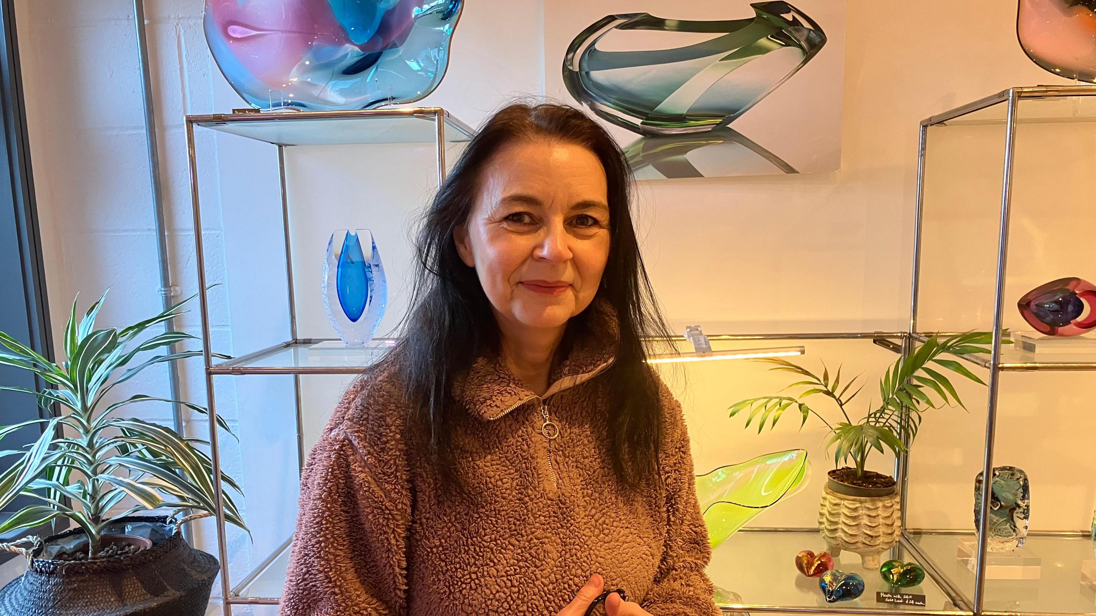 Nicola Williams who owns Kinki Glass is standing smiling into the camera with some glass art work sitting behind her on shelves 