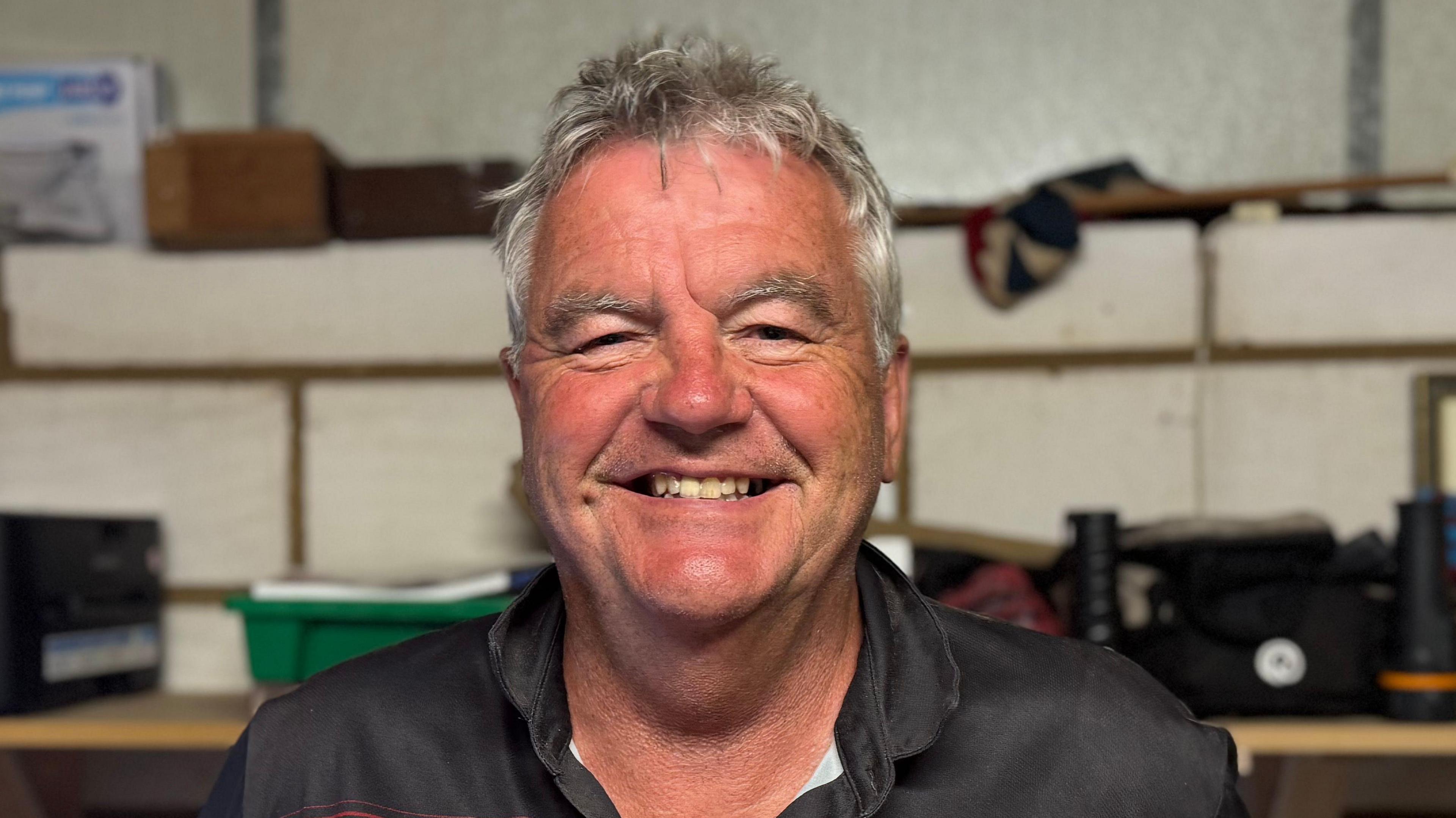 A head and shoulders image of Michael Hancock. He is wearing a black and red top and standing in front of a shelf that has various items on it. He is smiling and looking directly into the camera.