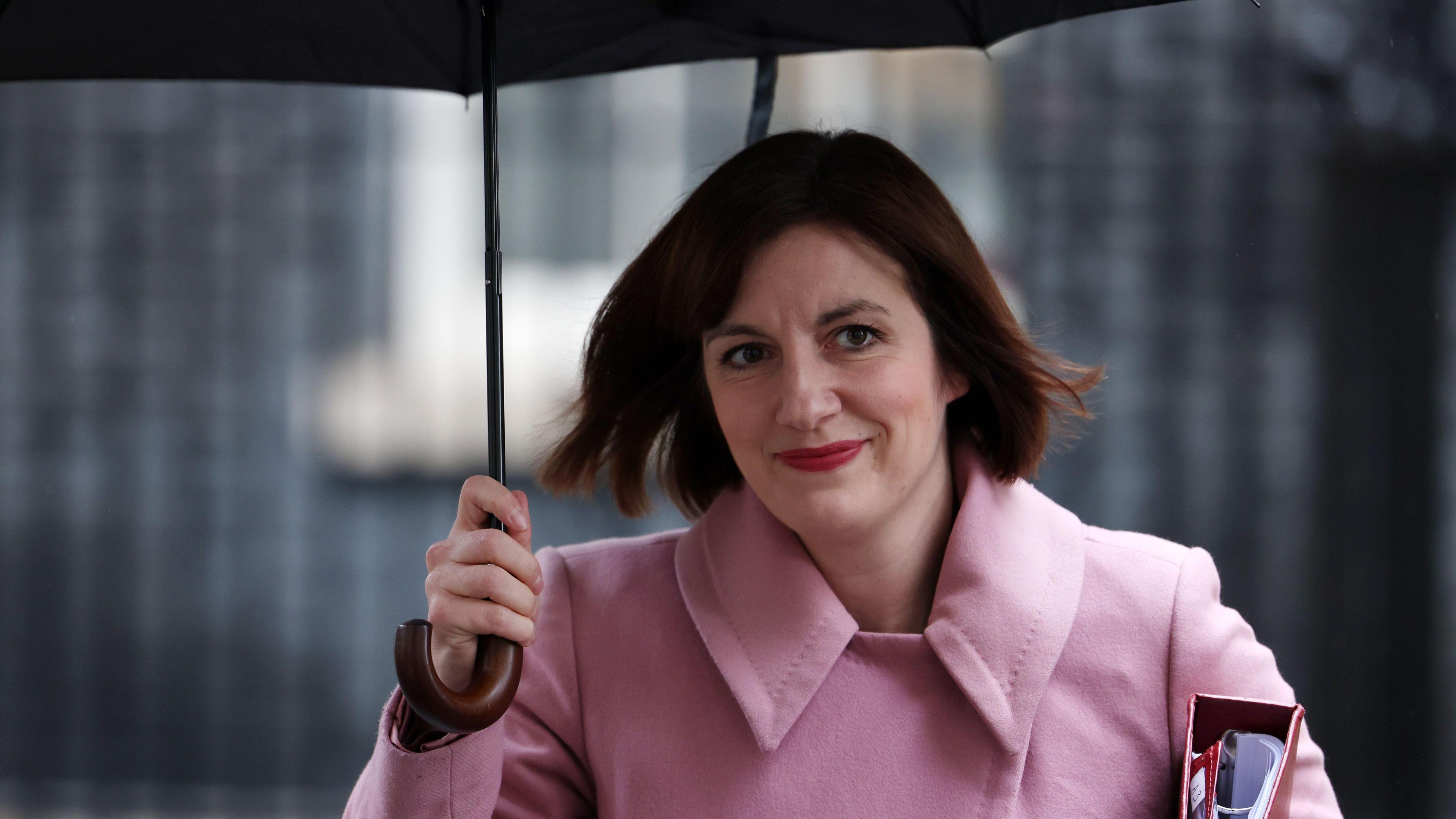 A woman in a pink coat holding an umbrella smiling at the camera