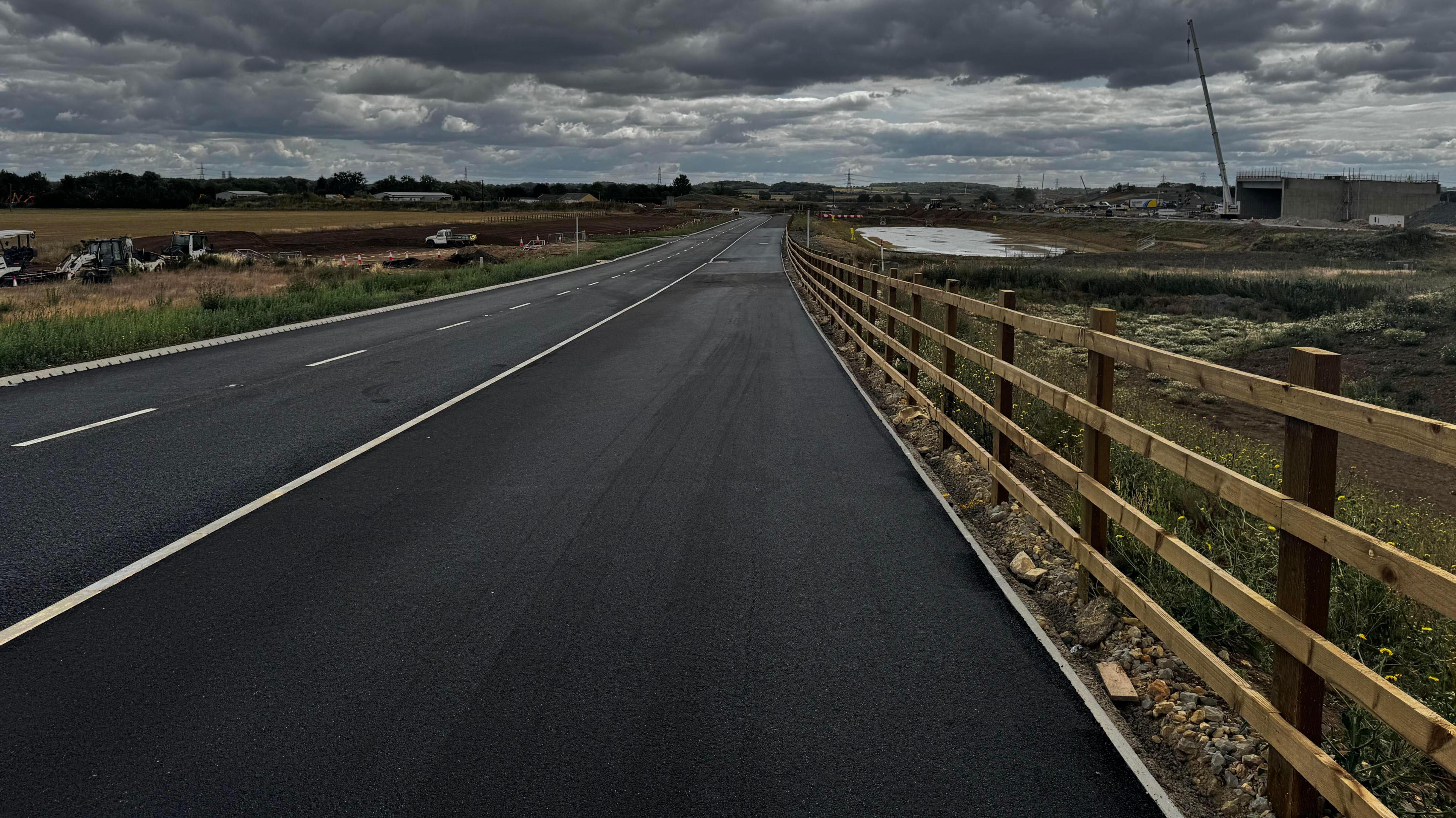 A new section of a new road, showing freshly laid tarmac, a wooden fence, along the righthand side, white markings on the road and machinery in the distance, including a large crane.
