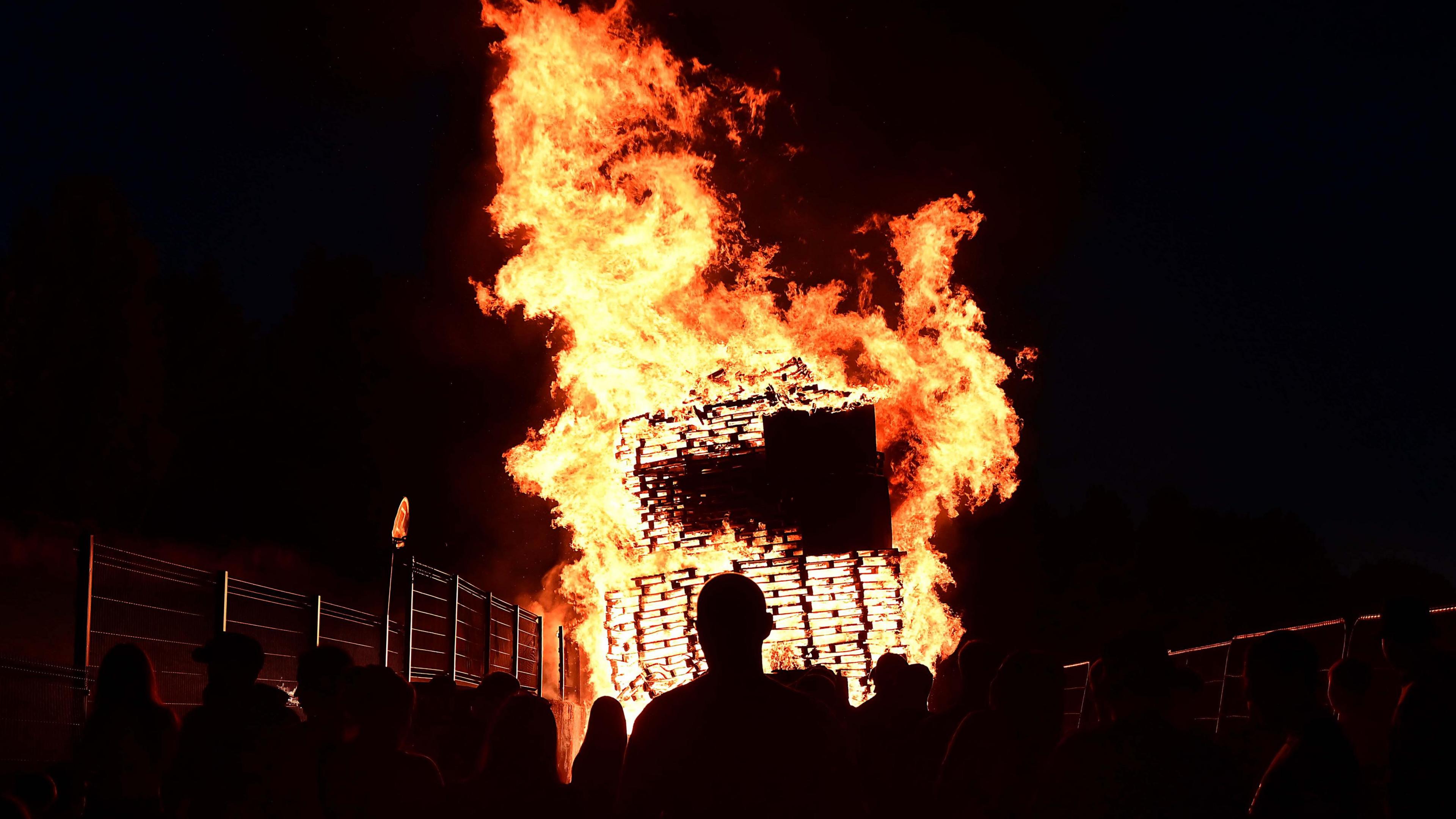 A bonfire goes up in flames, silhouetted against a black night's sky. In the foreground silhouetted figures can be seen watching it burn.