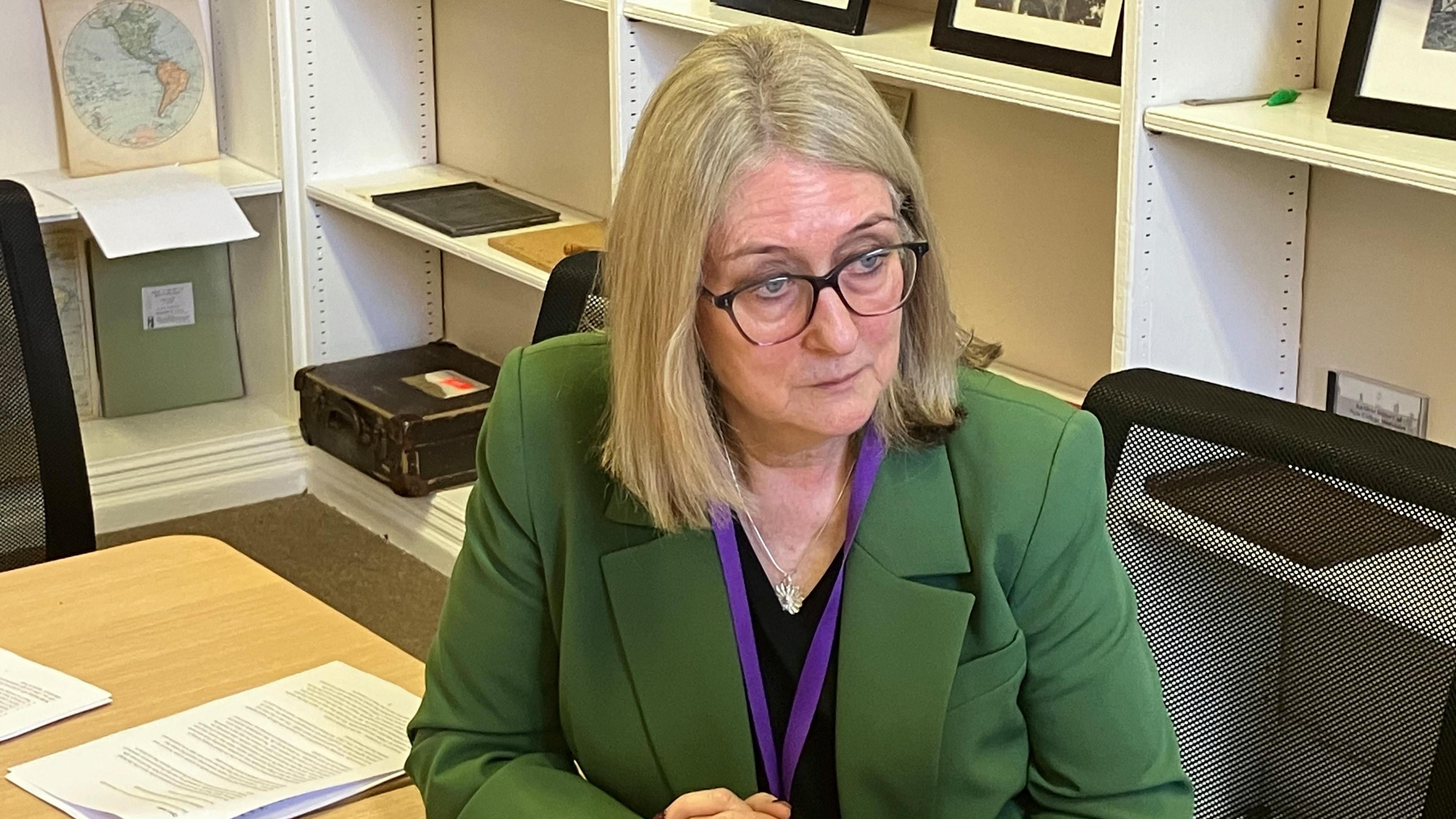 Jacqui Smith, the skills minister and government spokesperson for equalities, sits at a desk as she speaks to someone off-camera. She has shoulder-length grey-blonde hair and glasses and is wearing a black v-neck top underneath a green jacket.