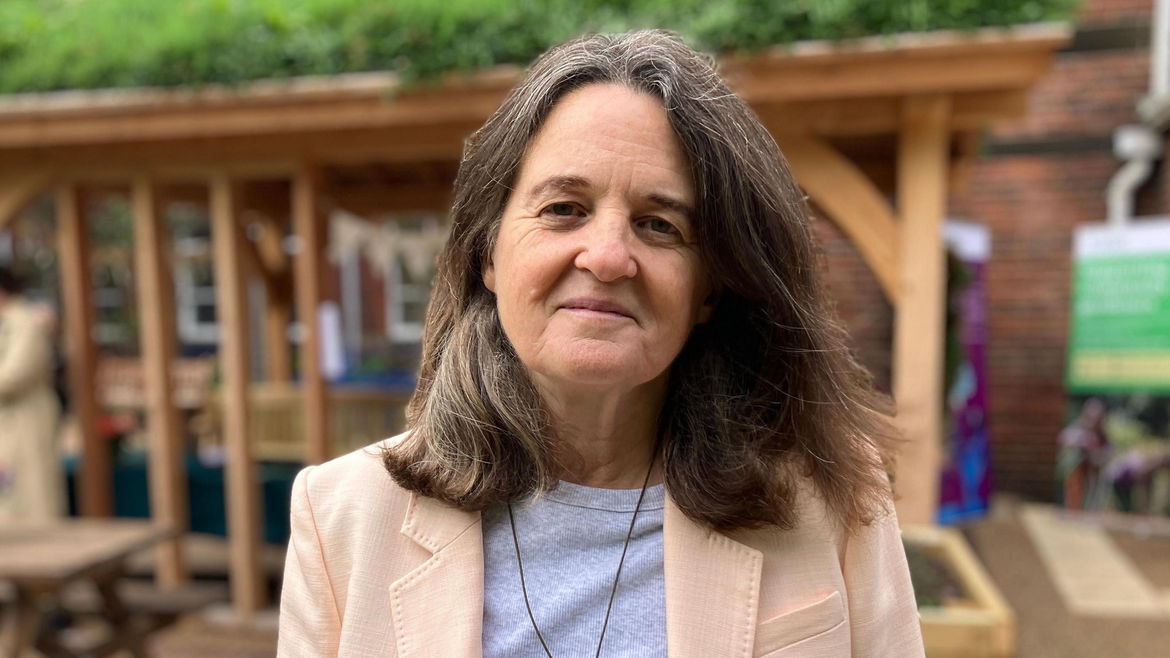 Clare Matterson, the RHS Director General, smiling at the camera from the garden. She is wearing a grey t shirt and pink jacket.
