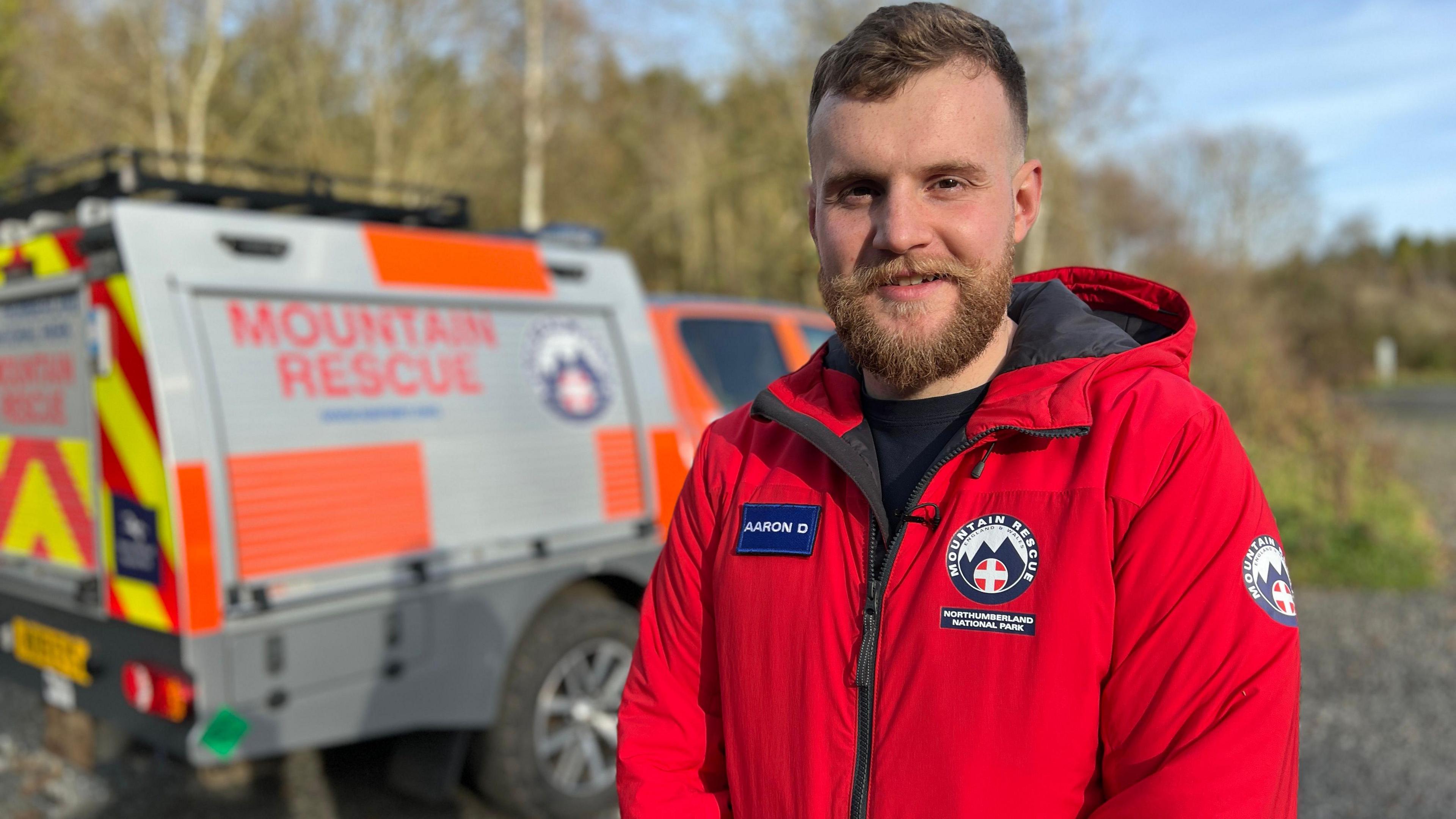 Aaron Duke, wearing red mountain rescue uniform overalls, stand in front of a 4x4 rescue vehicle. He is smiling looking into the camera. 