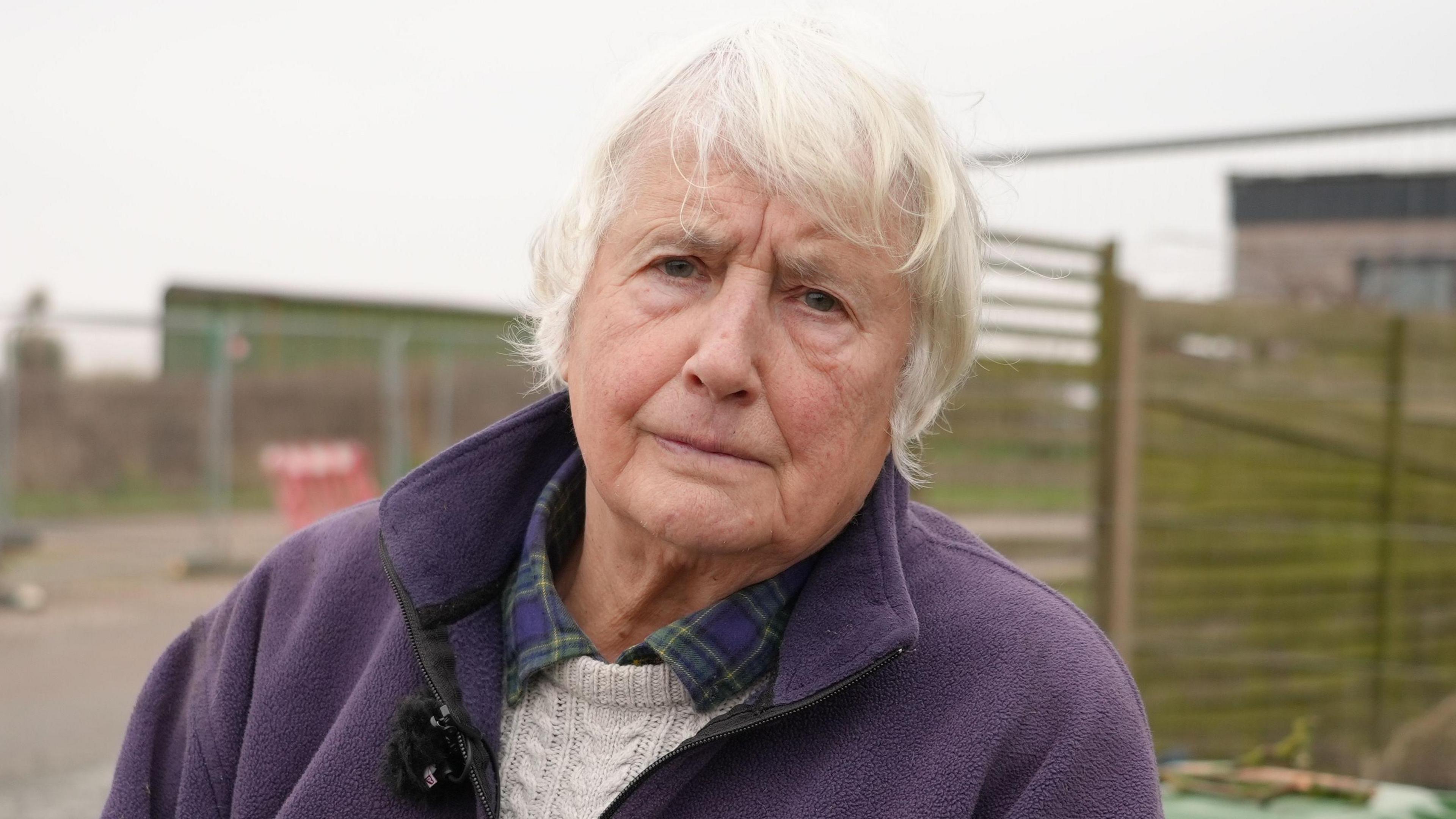 Bryony Nierop-Reading, a woman is standing outside near a cliff edge in Happisburgh. She is wearing a green and blue plaid shirt with a cream knitted jumper over the top and a purple fleece. She is looking directly at the camera.
