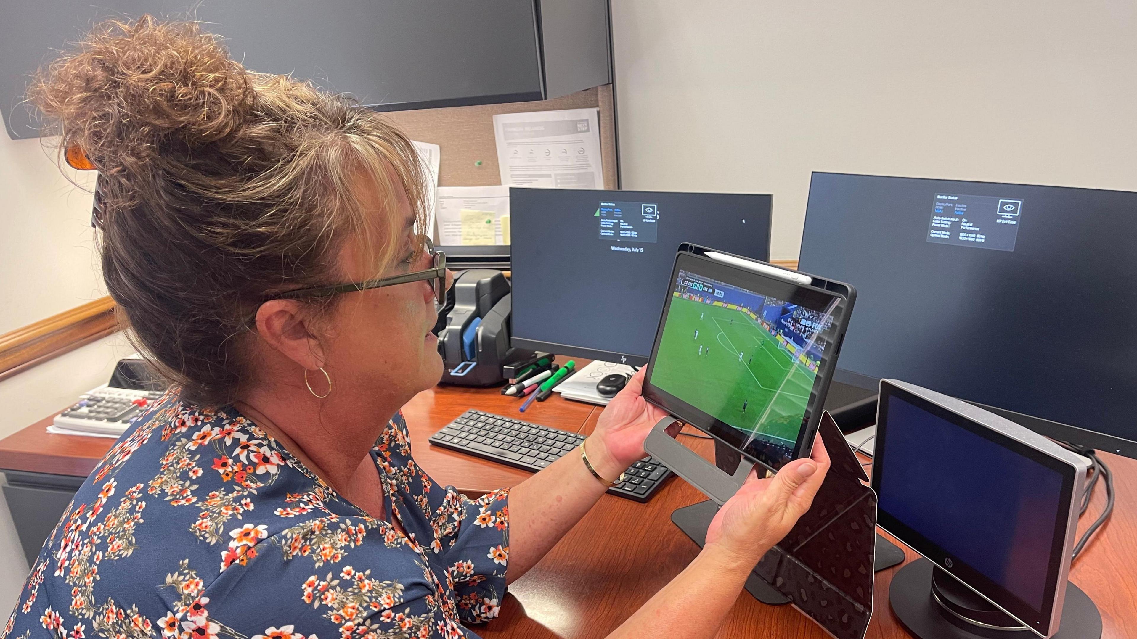 Heather Earhart, 54, wears a floral shirt and glasses, her curly blonde hair tied up, as she sits at her desk holding a tablet that's showing the England versus Argentina match